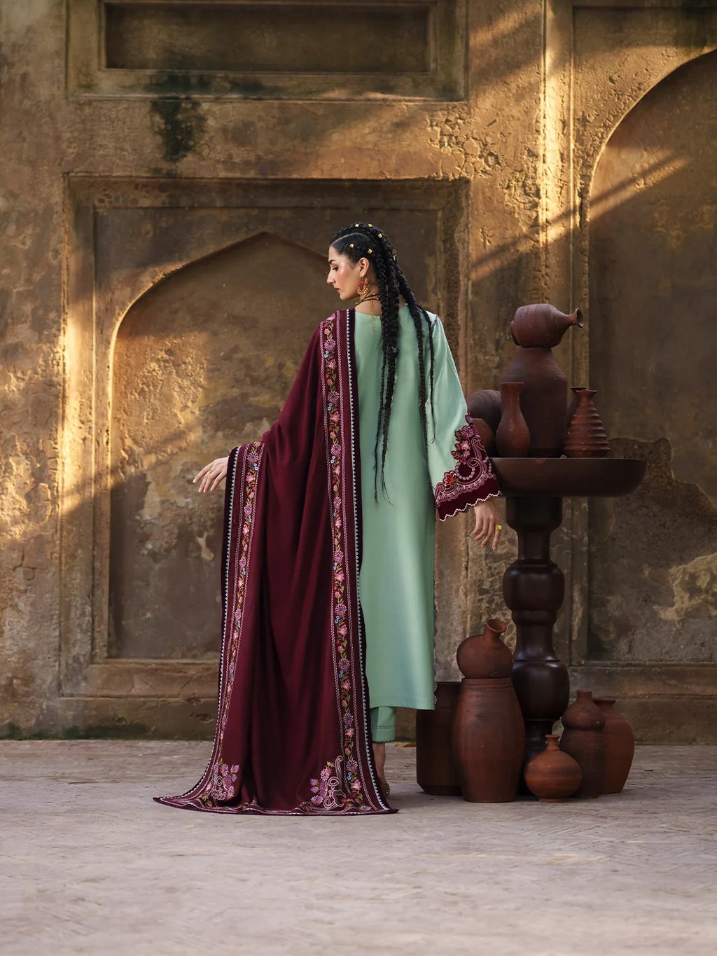A woman wearing a traditional outfit stands in profile in front of a textured stone wall with arches. She is wearing a mint green kurta with a deep burgundy dupatta featuring intricate floral embroidery.