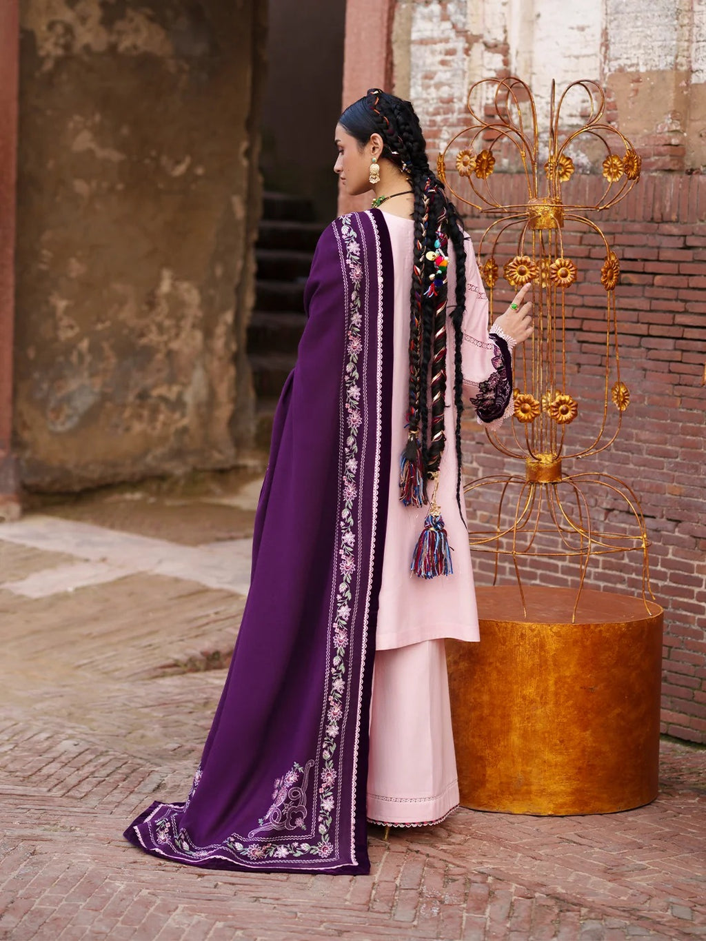 A woman wearing a traditional South Asian outfit with a light pink kurta and a deep purple dupatta with intricate silver embroidery, standing in an outdoor courtyard next to a decorative gold metal sculpture.
