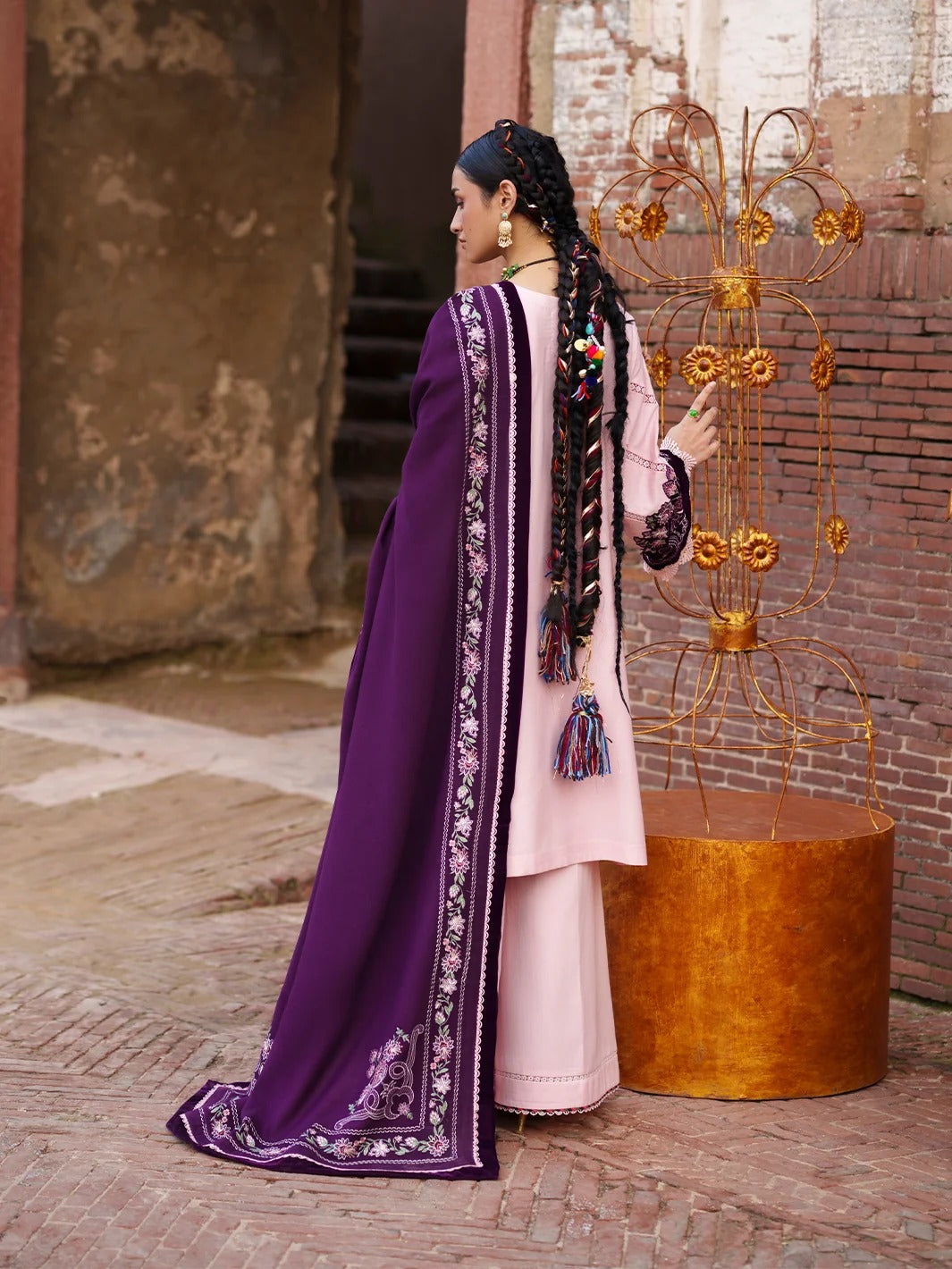 A woman wearing a traditional South Asian outfit with a light pink kurta and a deep purple dupatta with intricate silver embroidery, standing in an outdoor courtyard next to a decorative gold metal sculpture.