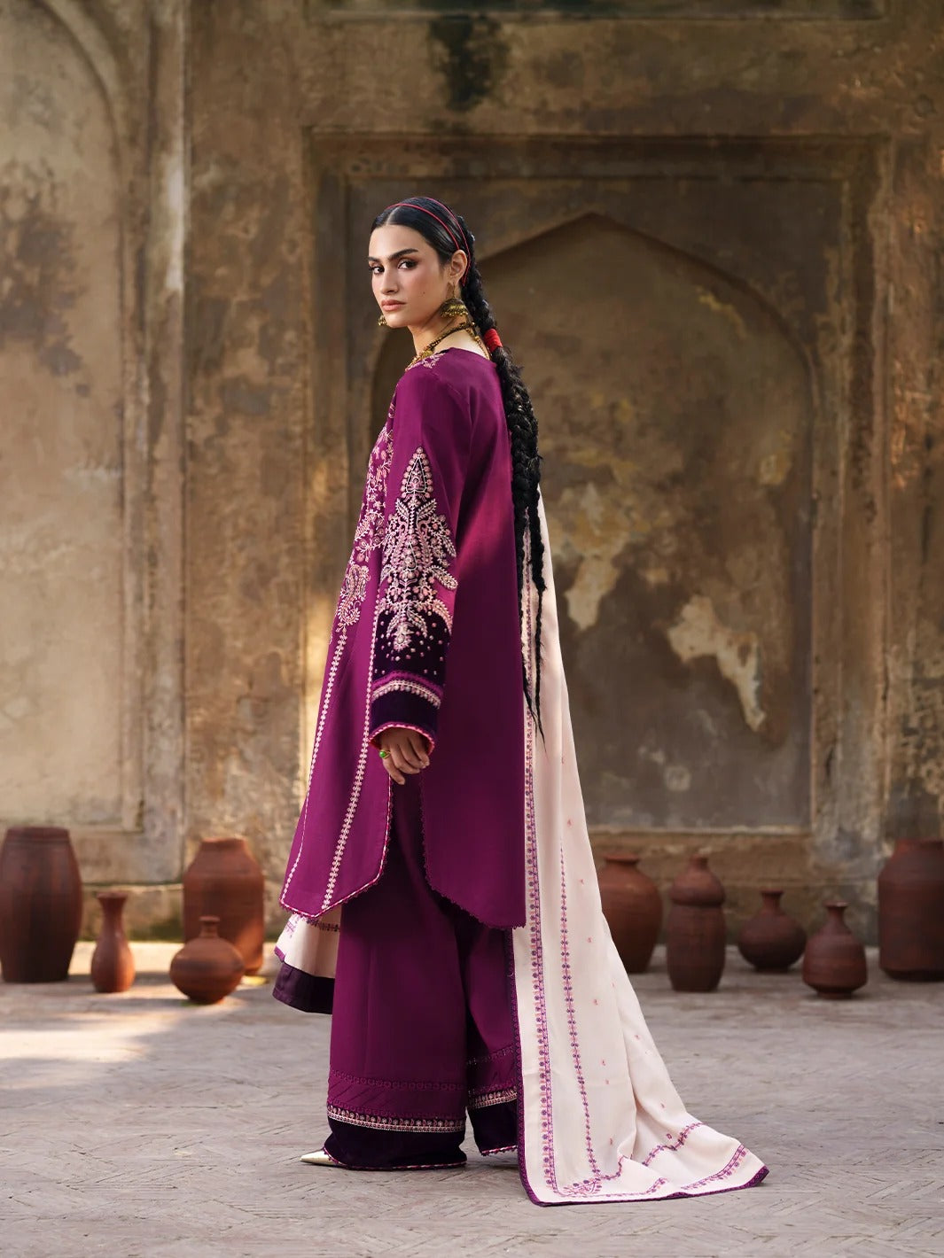 A woman models a traditional South Asian outfit consisting of a deep purple kurta and wide-leg trousers with intricate embroidery, paired with a cream-colored dupatta featuring pink border detailing, set against a rustic stone wall with terracotta pots.
