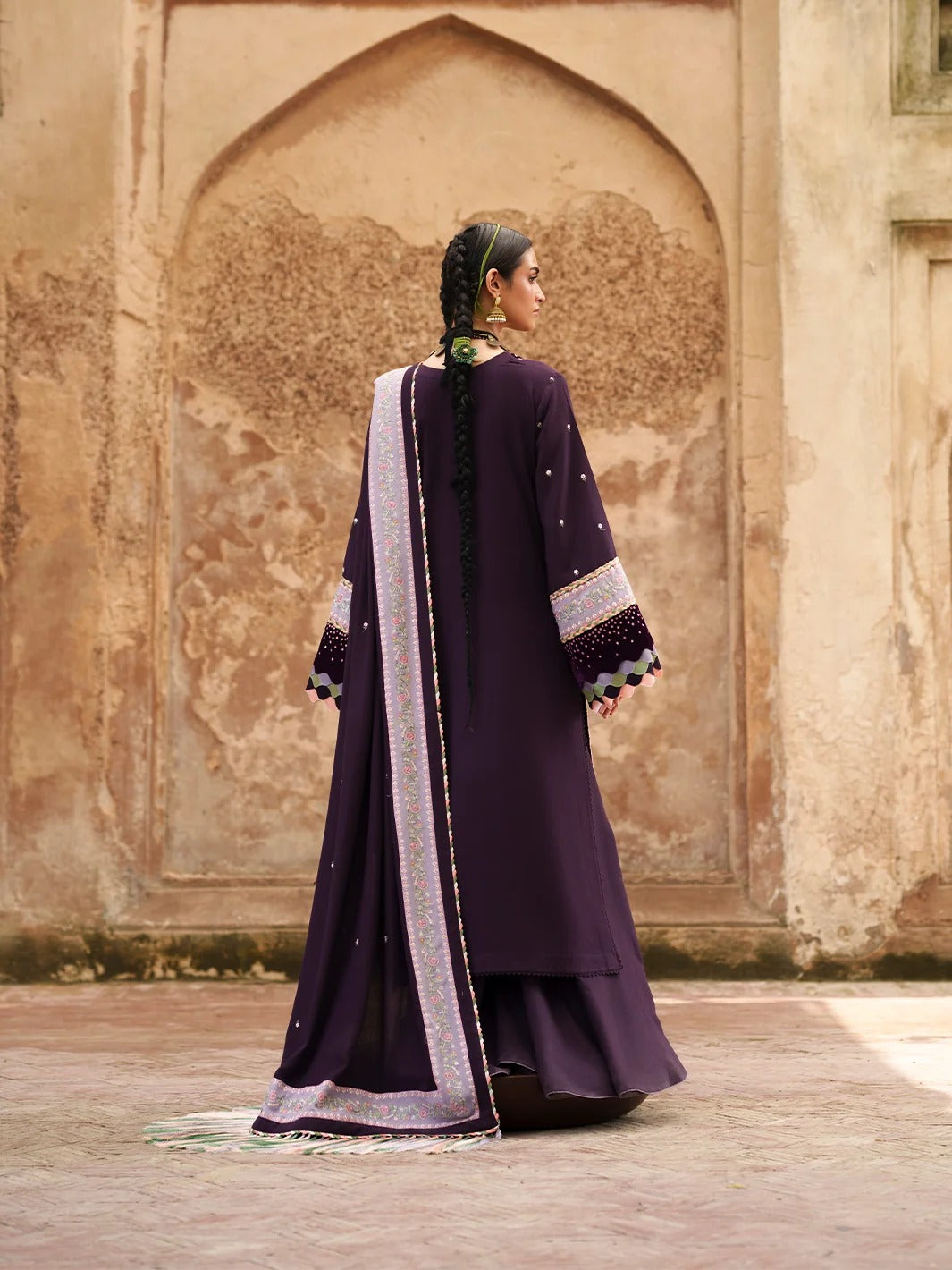 A woman wearing a traditional dark purple Anarkali kurta with a matching dupatta is photographed from behind, standing in front of a textured beige stone wall with an arched niche.