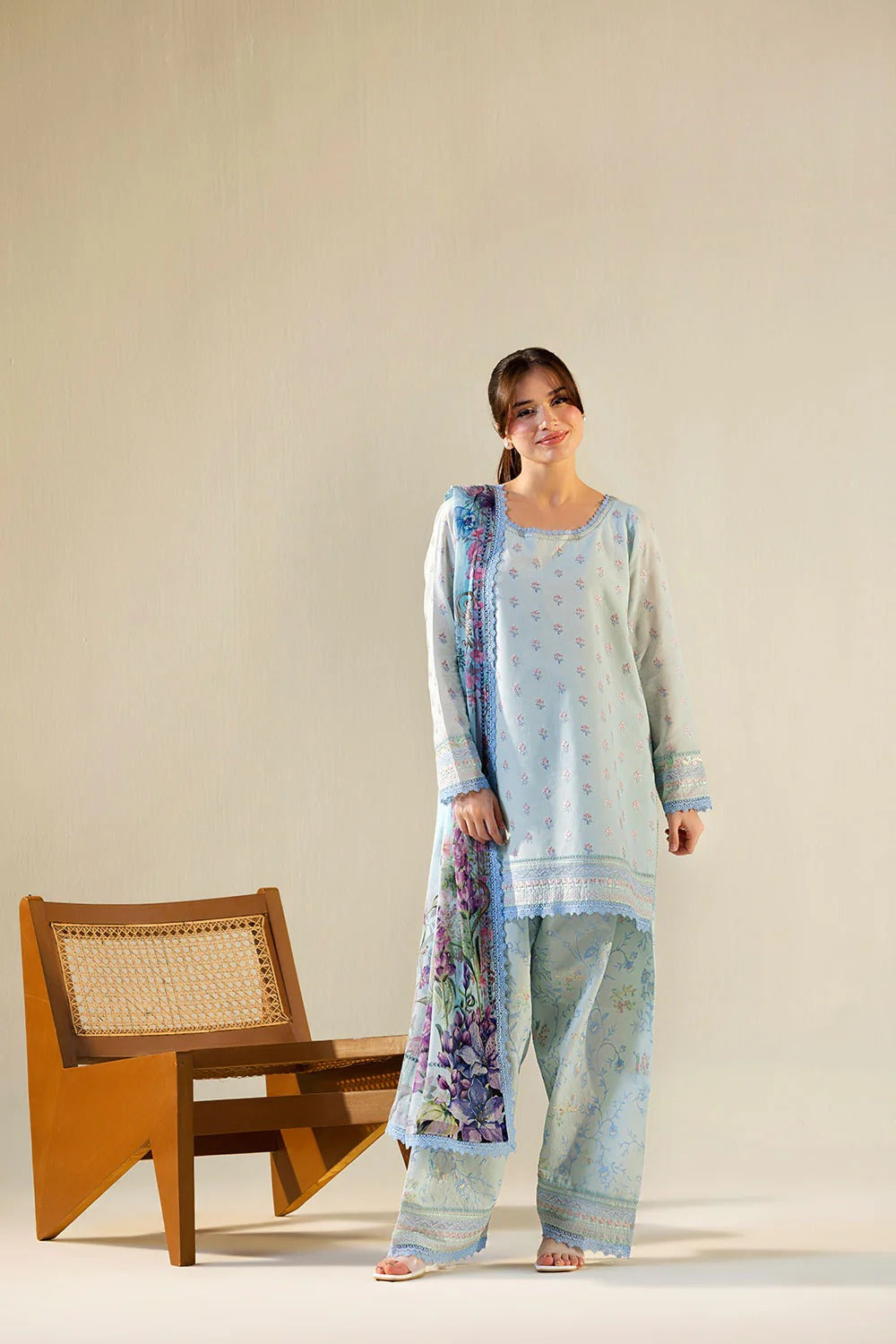 Woman in a light blue traditional outfit with a floral shawl standing next to a wooden chair on a beige background
