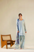 Woman in a light blue traditional outfit with a floral shawl standing next to a wooden chair on a beige background
