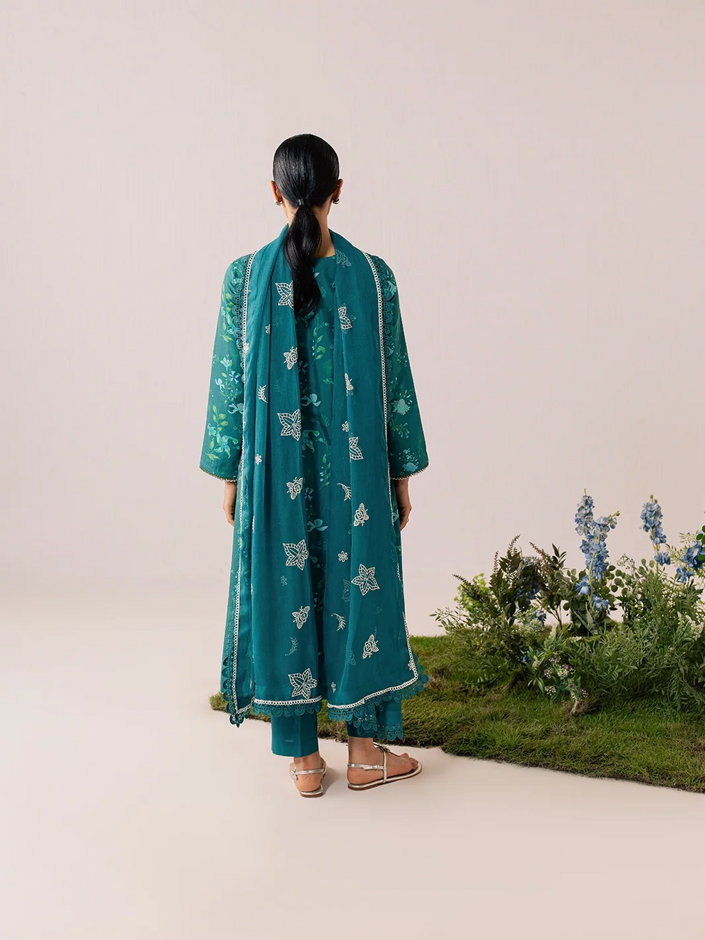 A woman stands with her back to the camera wearing a teal green traditional outfit with white printed motifs and a matching dupatta. She is photographed in a studio setting with a beige background and a small decorative plant arrangement on the right.