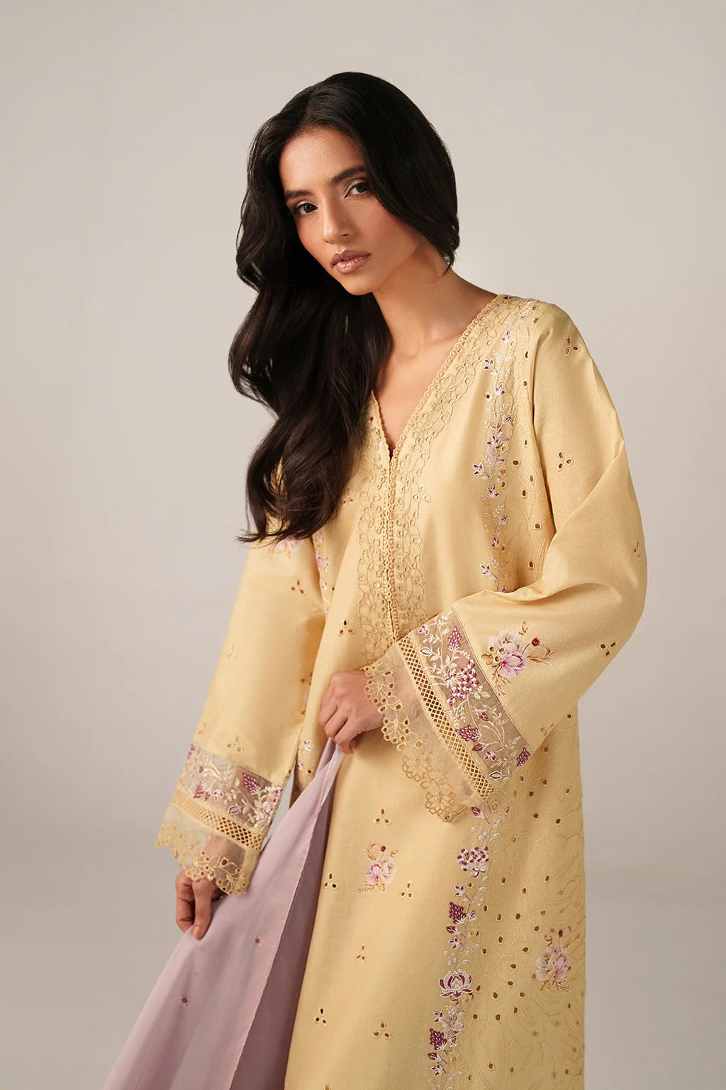Woman wearing a yellow embroidered dress holding a pink scarf against a neutral background