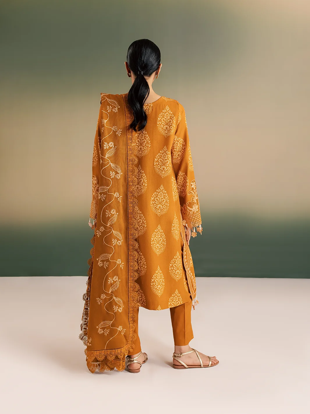 A woman stands with her back to the camera wearing a mustard yellow traditional outfit with beige floral embroidery, matching pants, and beige sandals, set against a soft green and beige studio background.