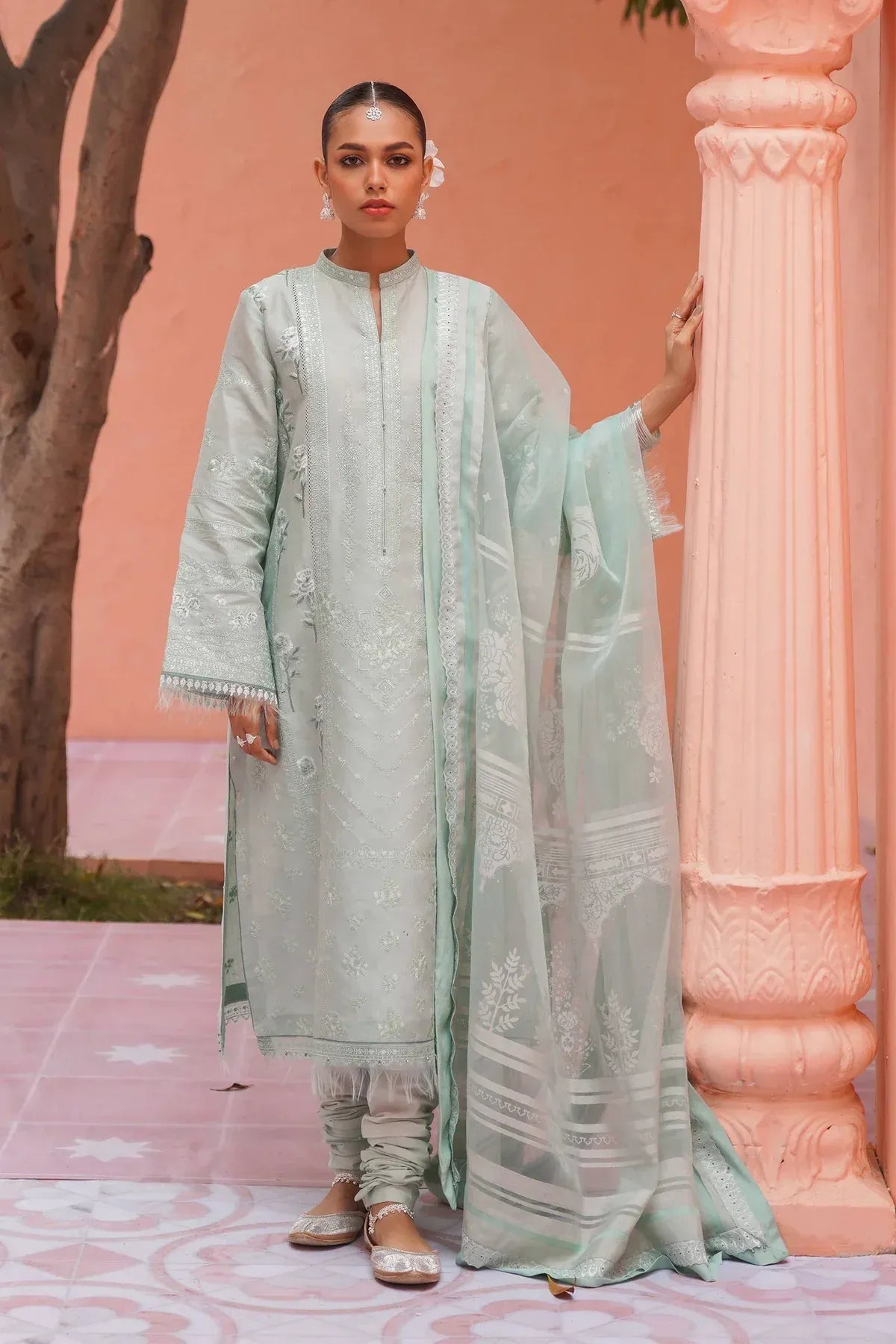 A woman models a mint green traditional South Asian outfit with matching embroidered dupatta and light gray trousers, standing in front of a pink architectural backdrop with a tree trunk on the left.