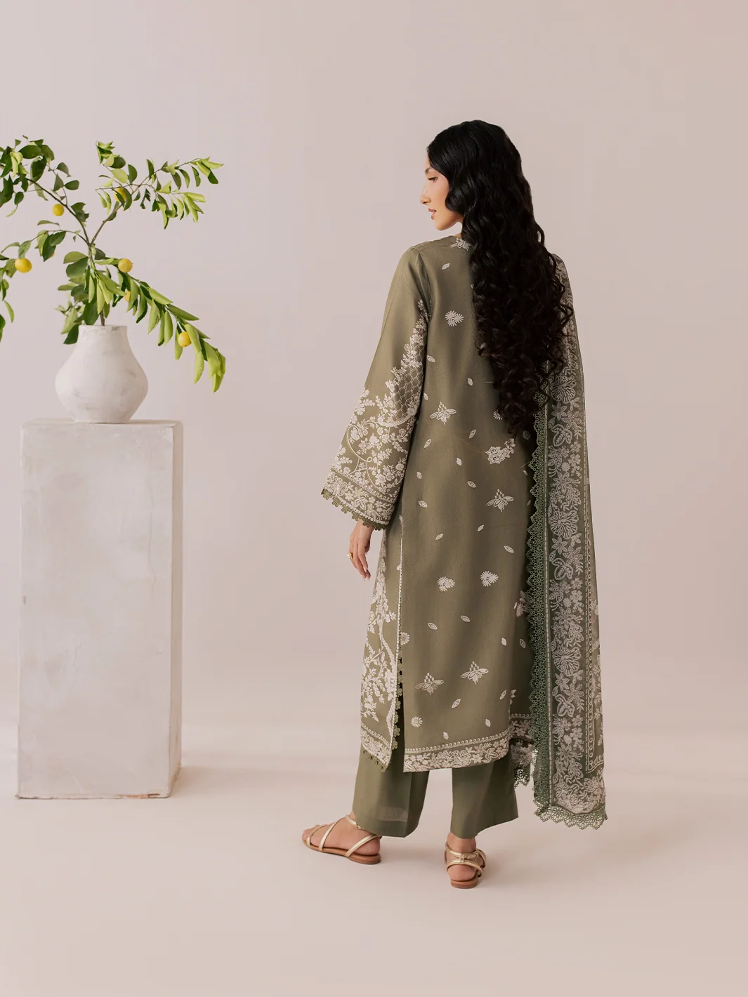 A woman stands with her back to the camera wearing an olive green traditional outfit with white floral embroidery and a matching embroidered dupatta. She is posed next to a white pedestal with a green leafy branch in a vase, against a solid light beige background.
