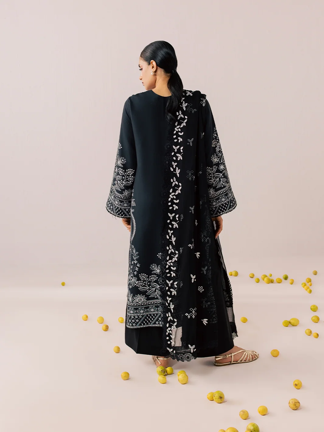 A woman stands with her back to the camera wearing a long black embroidered tunic or kurta with white floral and geometric patterns. The setting is a minimalist studio with a light beige background and scattered yellow lemons on the floor.