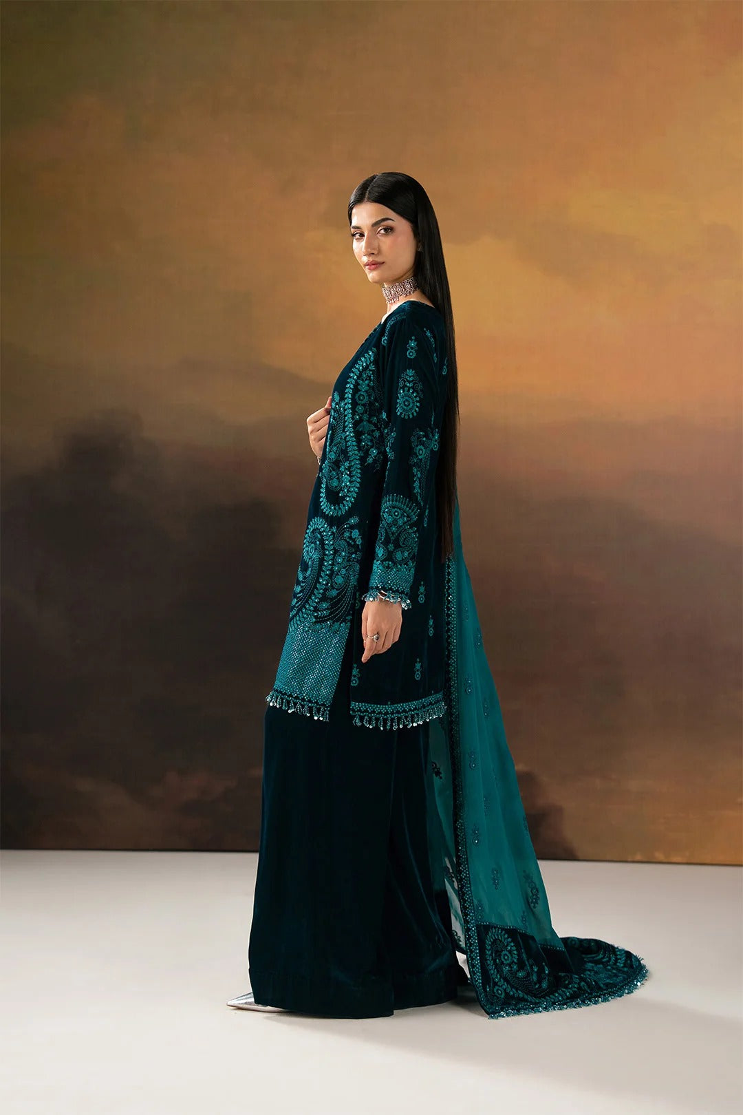 A woman modeling a dark teal embroidered traditional outfit with a long dupatta, standing against a warm brown and gold studio backdrop.