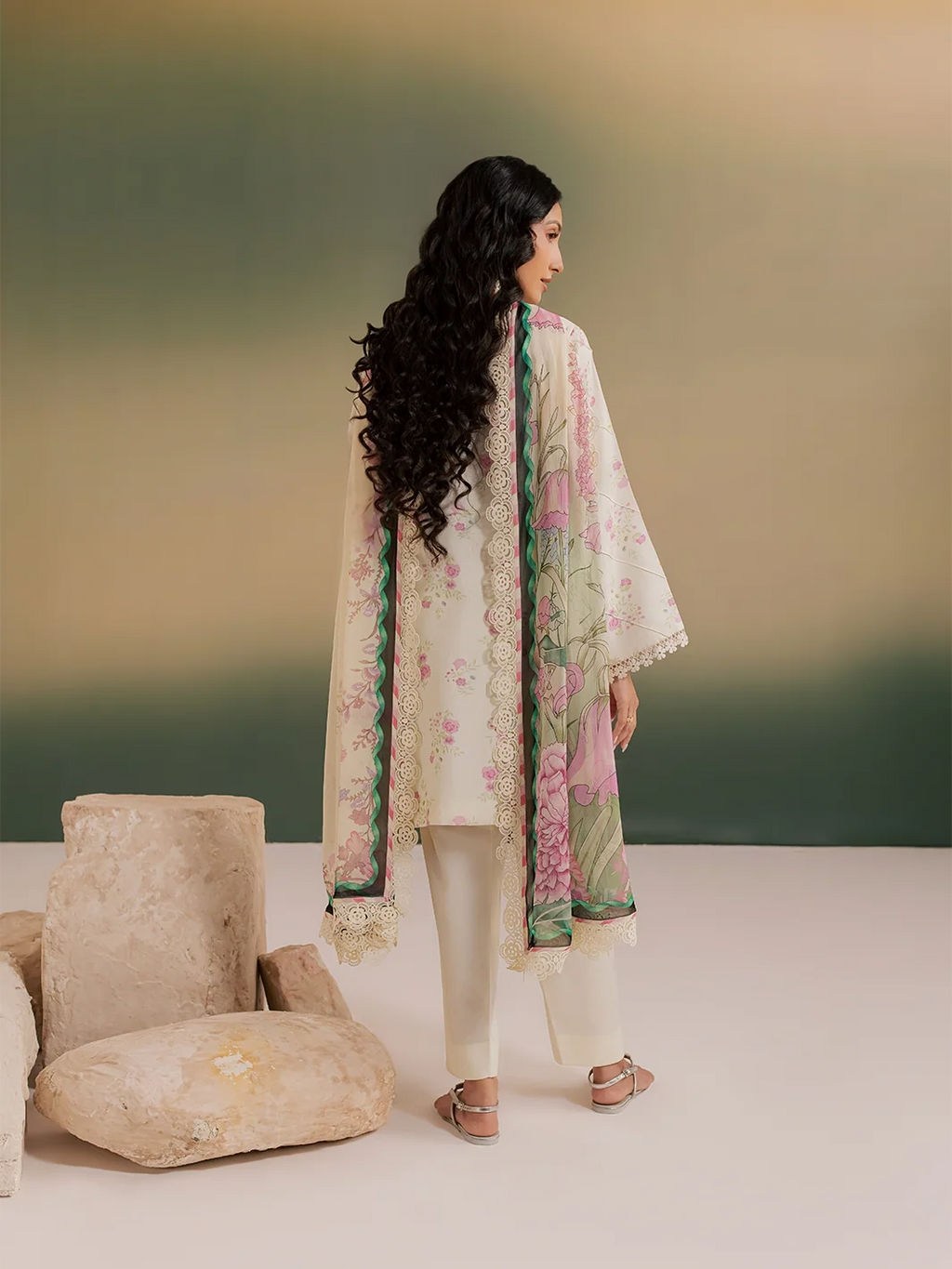 A woman stands with her back to the camera wearing a three-piece traditional outfit. The set features an off-white kurta with green and pink floral embroidery and lace trim, paired with matching off-white trousers and a sheer off-white dupatta with green and pink floral patterns and lace borders.
