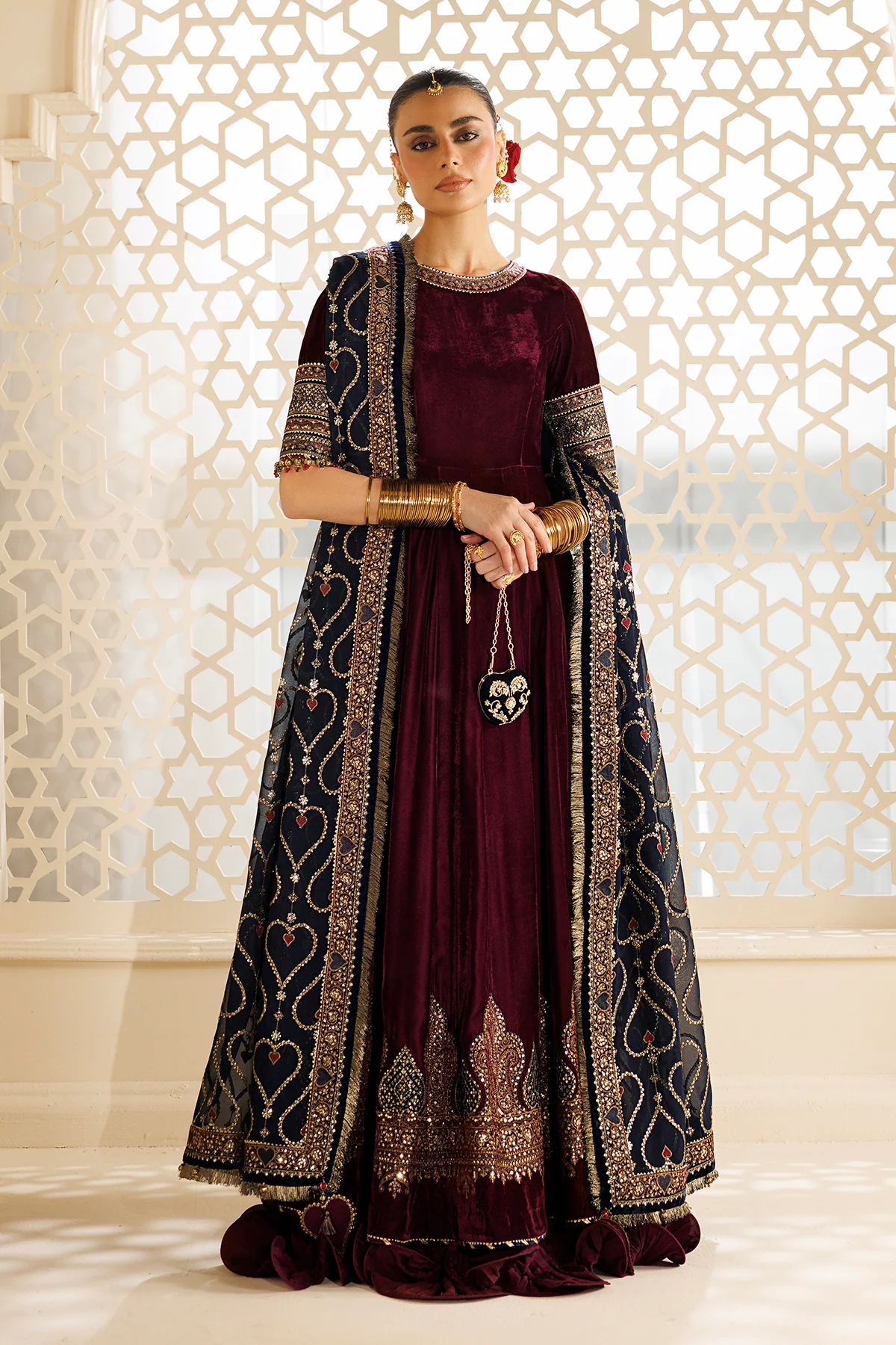 A woman models a traditional Indian outfit consisting of a deep maroon velvet anarkali dress with gold embroidery, paired with a matching dark navy blue dupatta featuring intricate gold motifs and borders. She is standing in front of a decorative beige geometric screen.