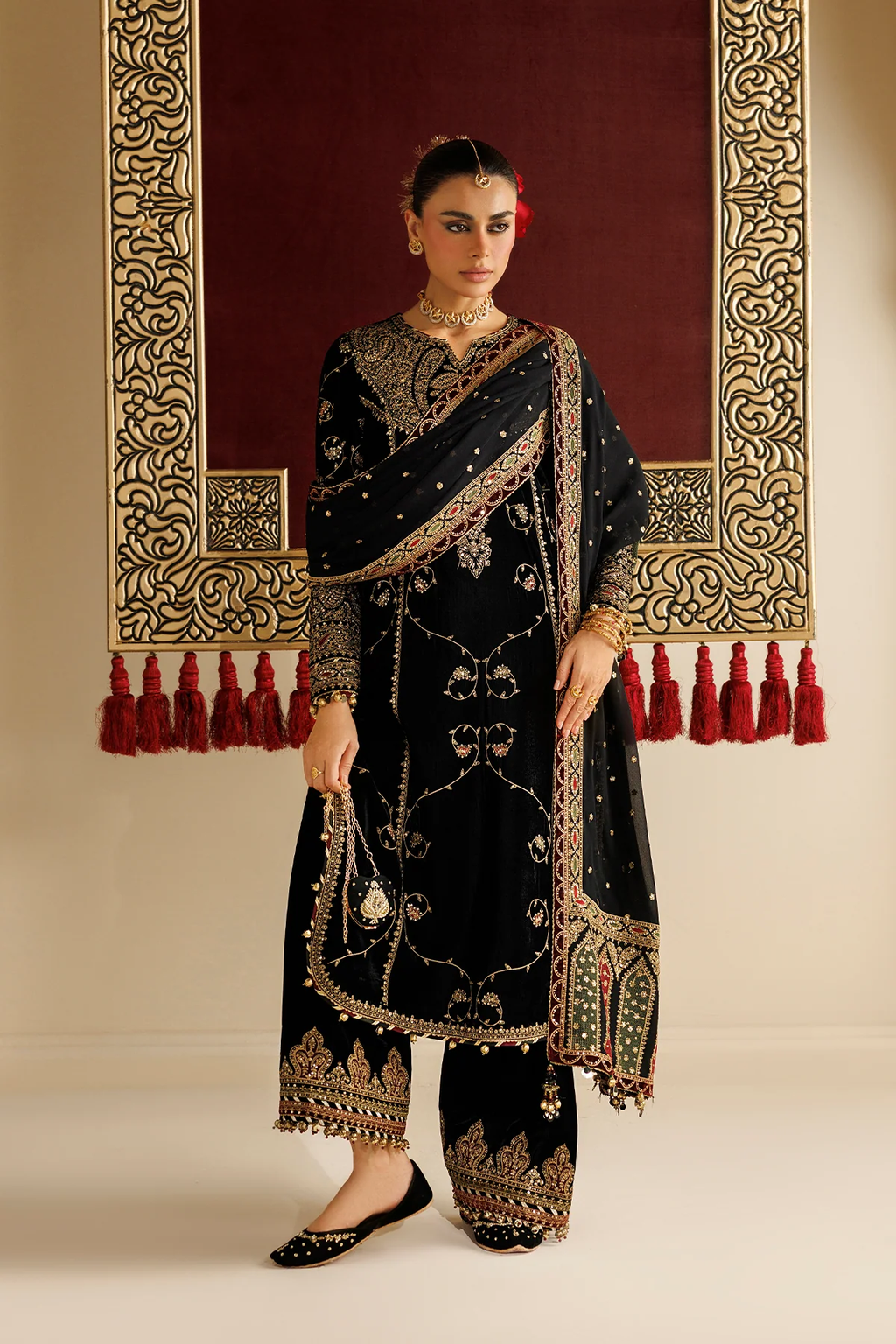 A woman models a black traditional ethnic outfit with extensive gold and silver embroidery, paired with a matching sheer dupatta, set against a warm beige wall with a large ornate red and gold hanging tapestry.