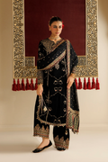 A woman models a black traditional ethnic outfit with extensive gold and silver embroidery, paired with a matching sheer dupatta, set against a warm beige wall with a large ornate red and gold hanging tapestry.