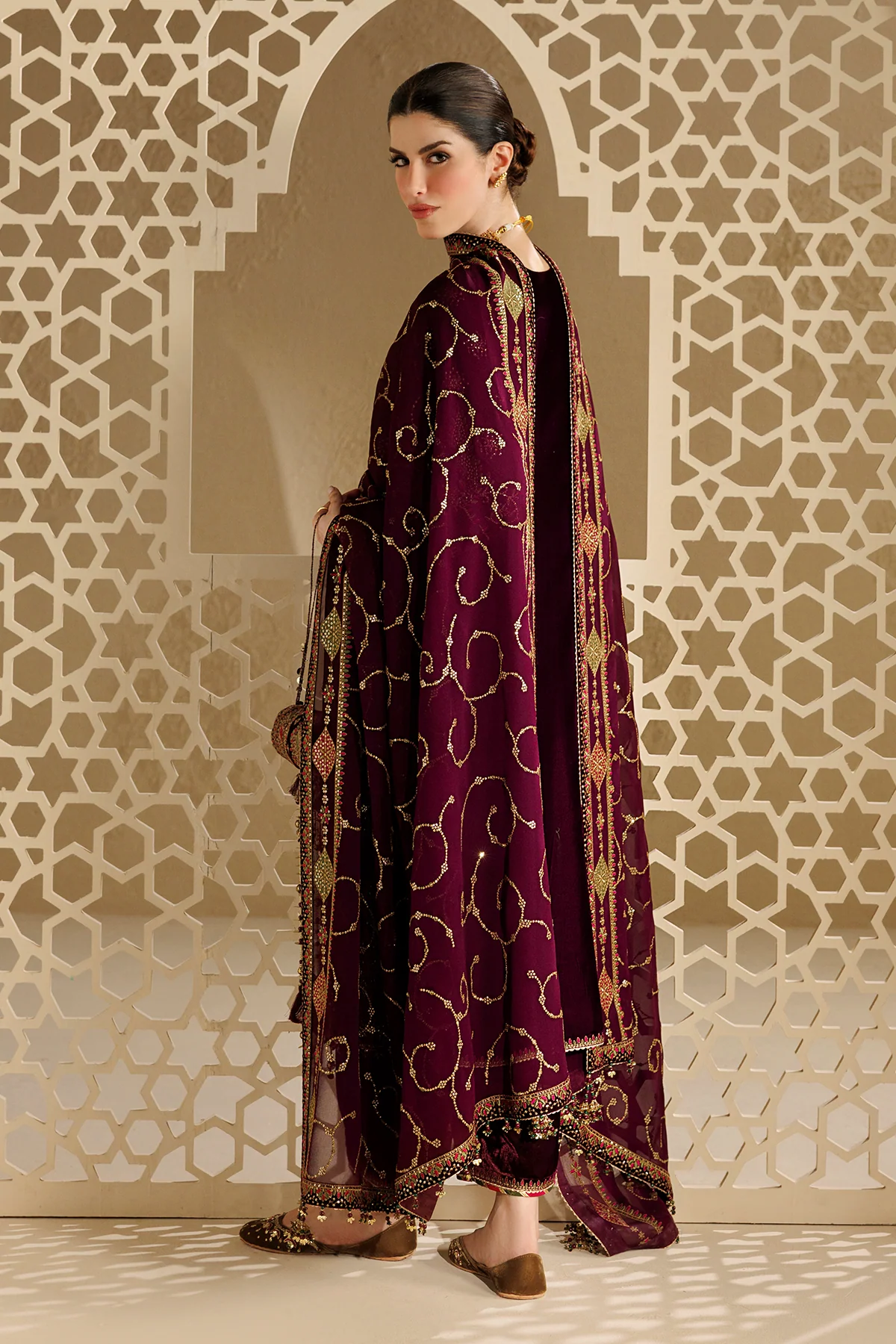 A woman models a deep purple velvet traditional South Asian outfit with intricate gold, pink, and green embroidery, standing in front of a beige archway with a geometric screen.
