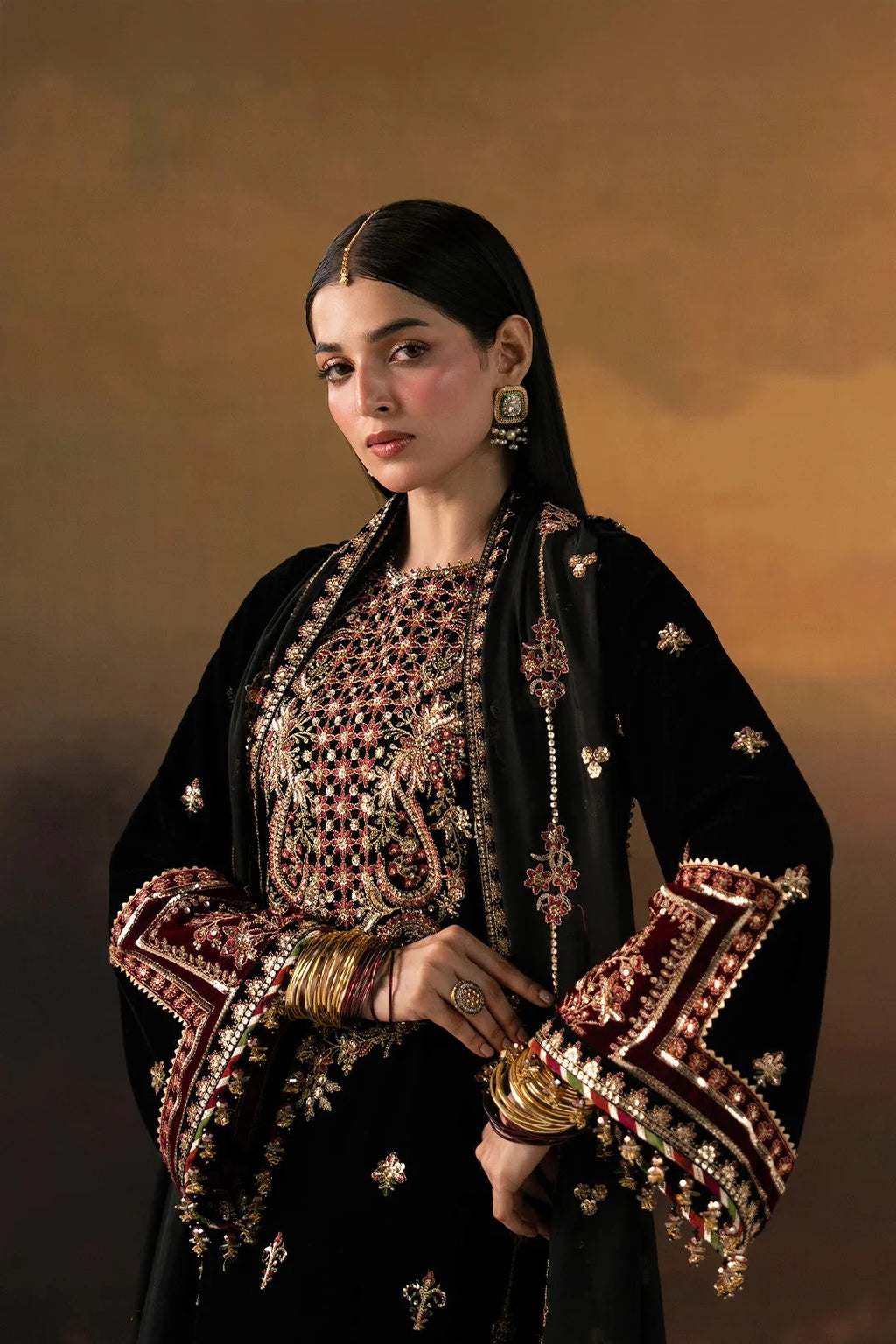 A woman wearing an ornate black traditional outfit with extensive gold, red, and white embroidery, styled with gold jewelry against a warm brown studio background.