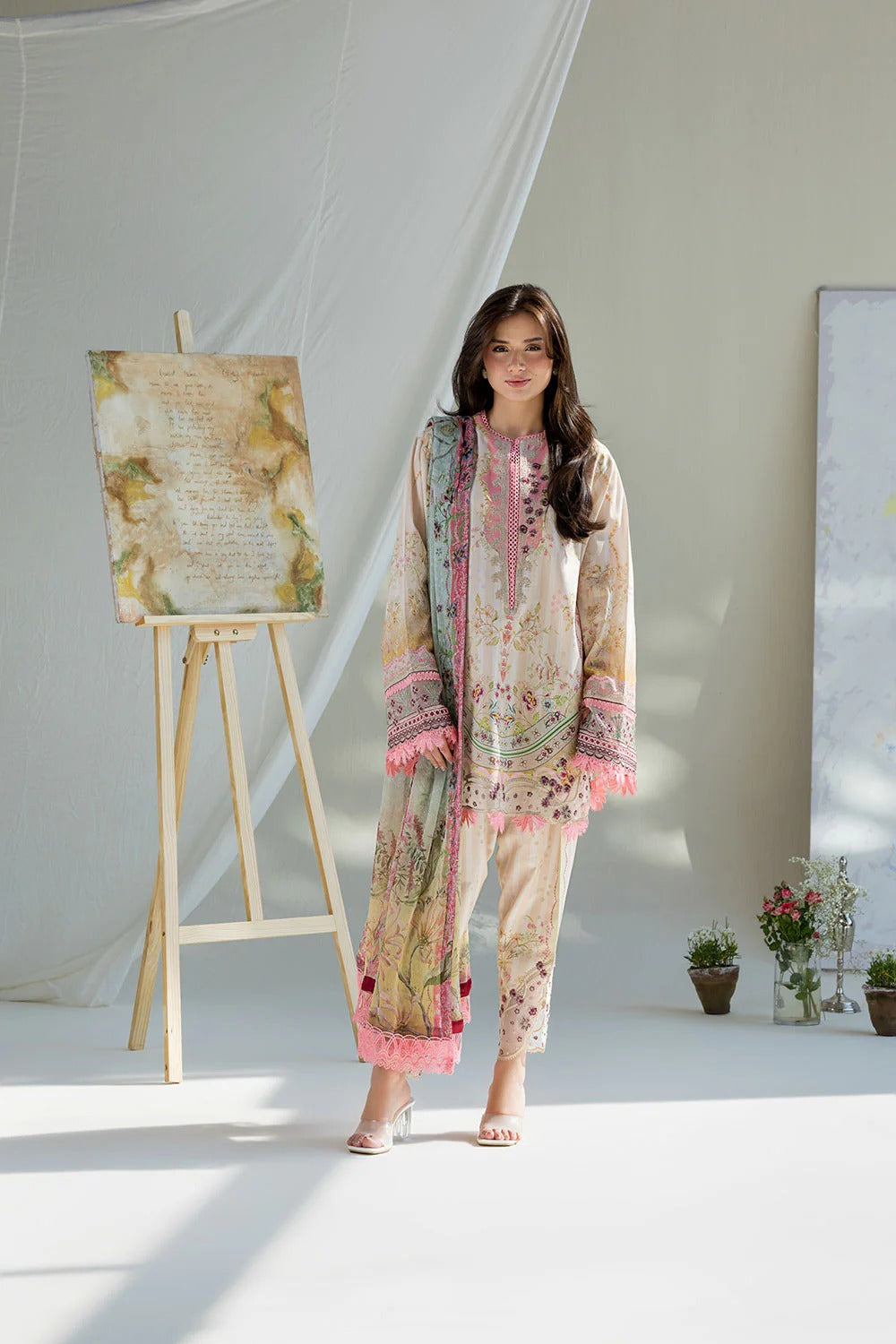 Woman in a floral dress standing next to an easel with a painting in a bright room.