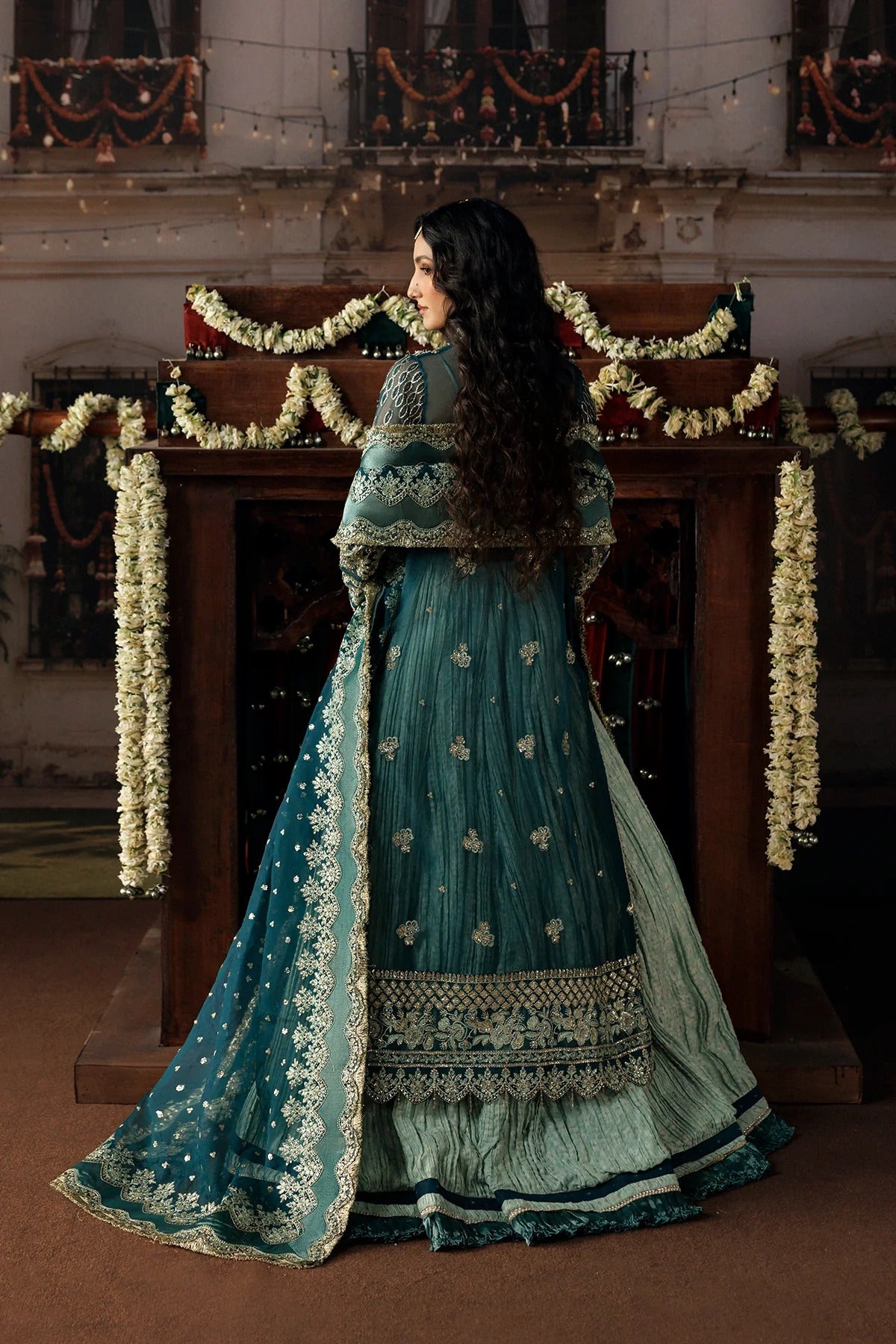 A woman stands with her back to the camera wearing a traditional teal green and gold embroidered Anarkali gown in an ornate indoor setting decorated with white flower garlands.