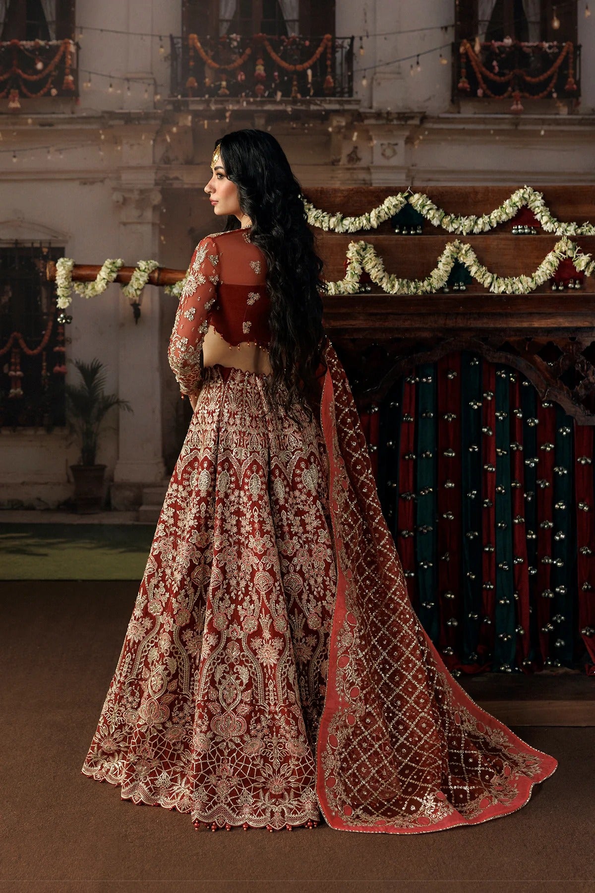 A woman wearing an ornate red and gold embroidered traditional Indian lehenga choli set, photographed in a decorated indoor venue with floral garlands and warm ambient lighting.