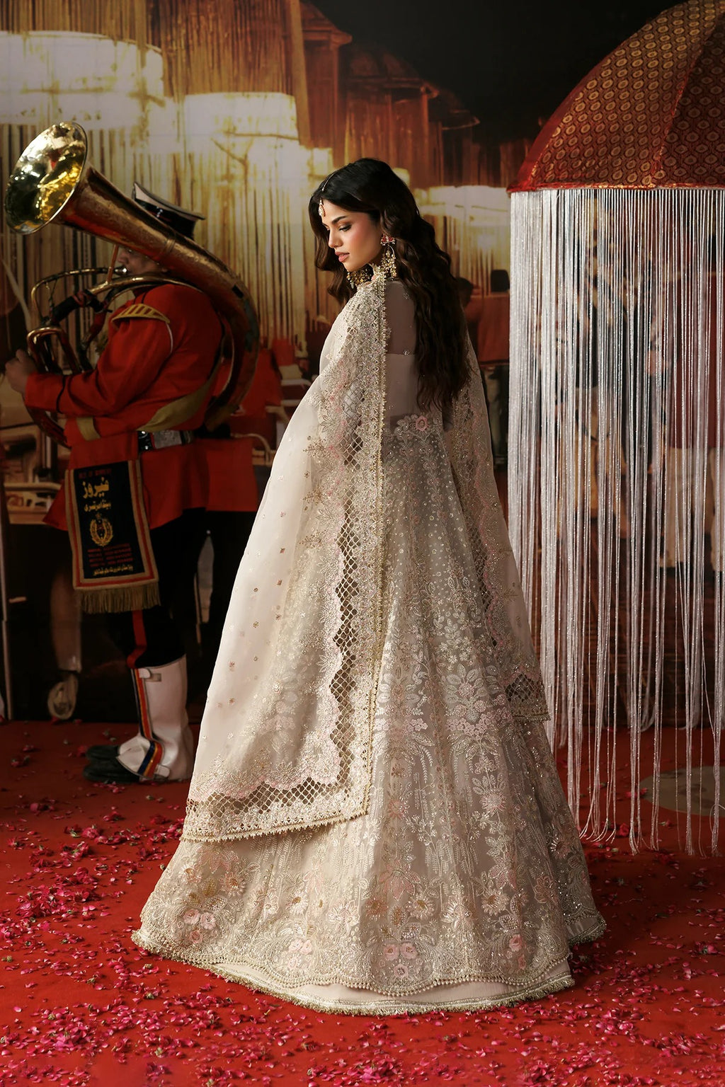 A woman wearing a traditional white embroidered lehenga choli with a sheer dupatta, standing on a red carpet with flower petals. A soldier in a red uniform with a brass instrument is visible in the background.