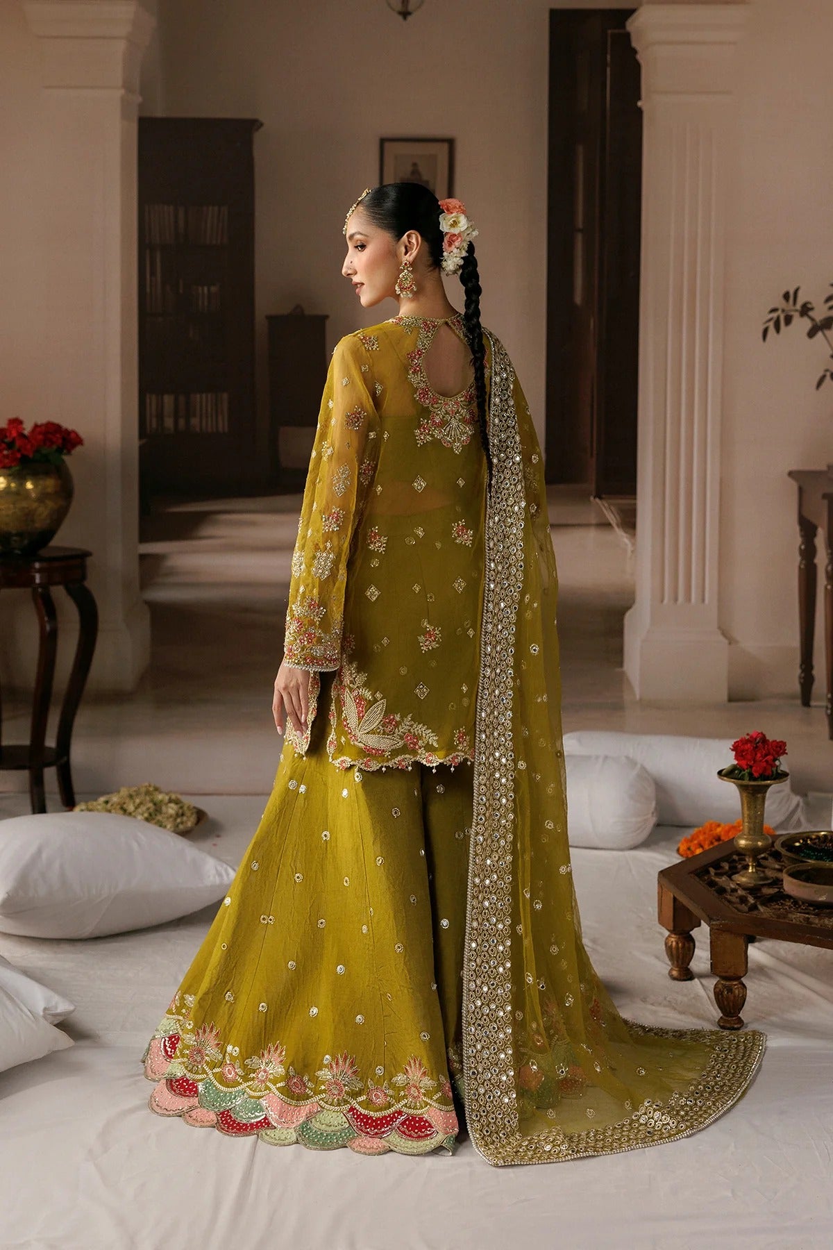 A woman wearing a traditional olive green embroidered ethnic outfit with a matching dupatta, standing in a decorated indoor room with white bedding and furniture.