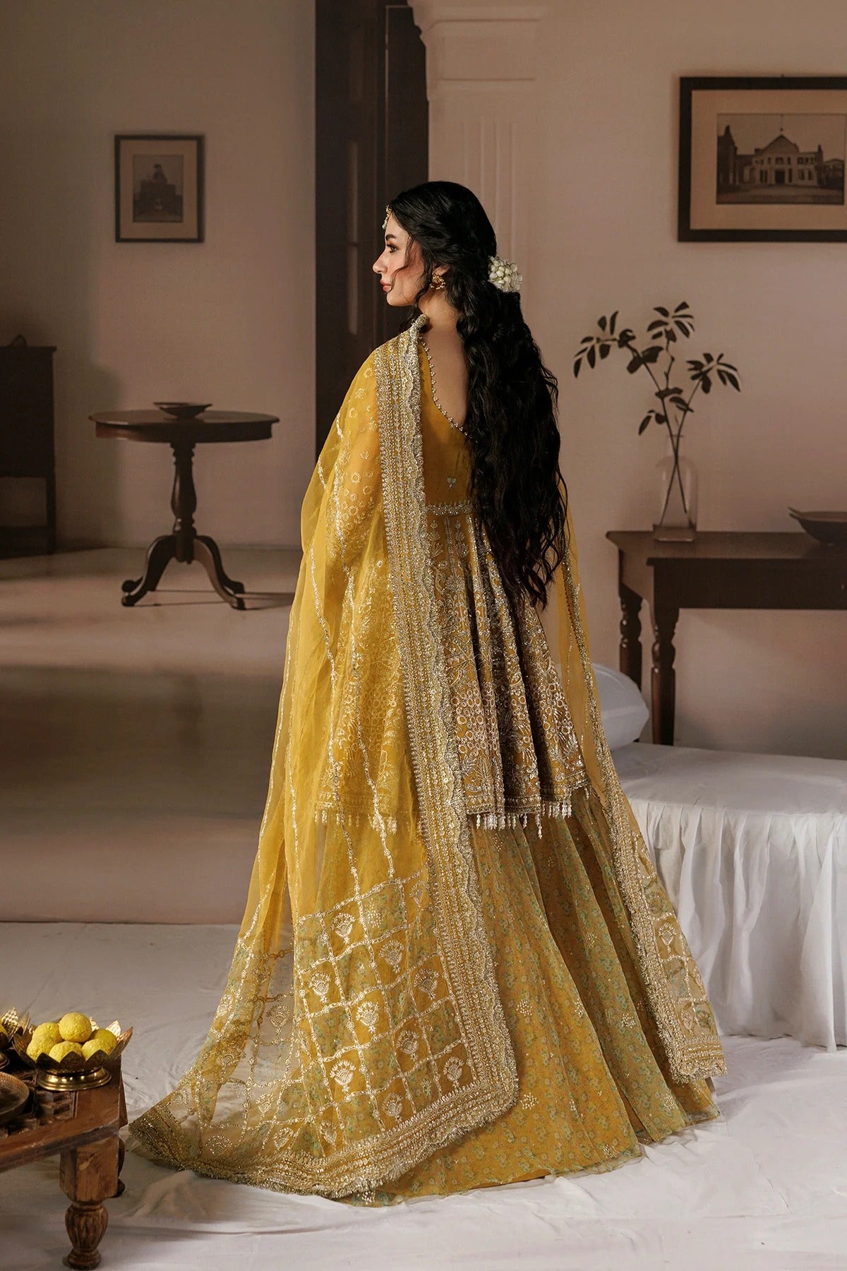 A woman wearing a traditional Indian lehenga choli in mustard yellow with intricate gold embroidery, standing in profile in a warm-toned room.