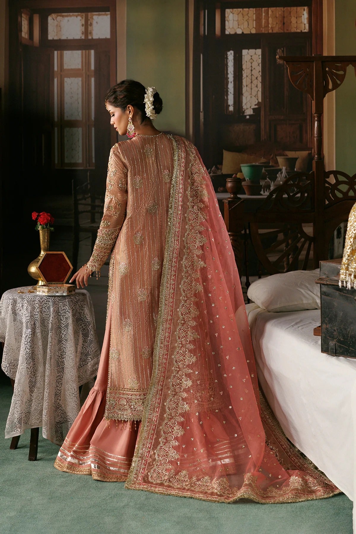 A woman wearing a peach-pink embroidered traditional outfit with a matching sheer dupatta, standing in a furnished room with wooden doors, a bed, and a small table with a red flower.