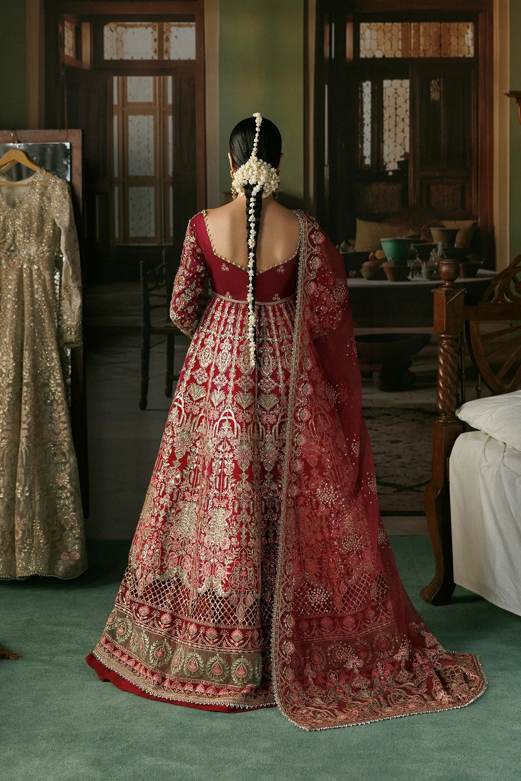 A woman stands with her back to the camera wearing an ornate red and gold embroidered traditional gown with a long draped dupatta in an indoor room. Another beige embroidered dress hangs on a hanger to the left.