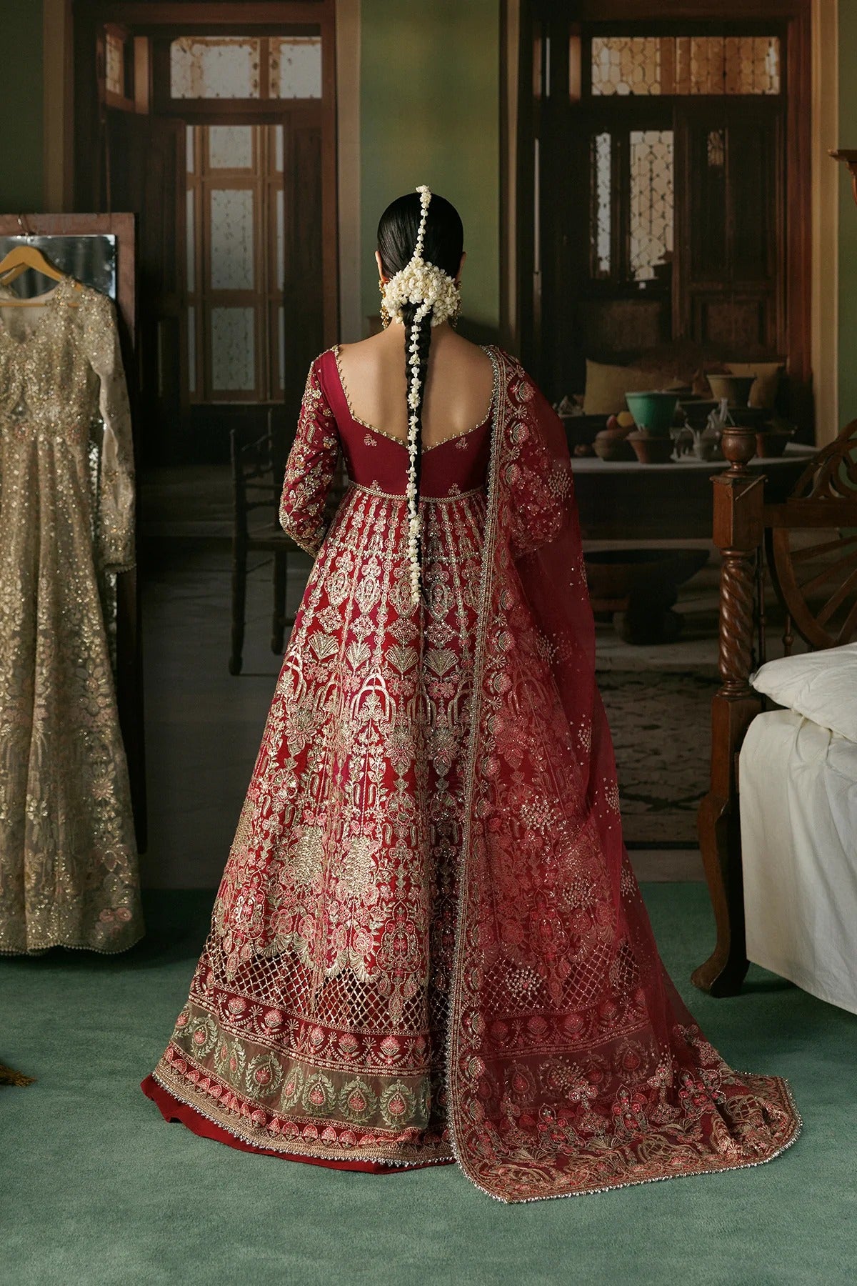 A woman stands with her back to the camera wearing an ornate red and gold embroidered traditional gown with a long draped dupatta in an indoor room. Another beige embroidered dress hangs on a hanger to the left.