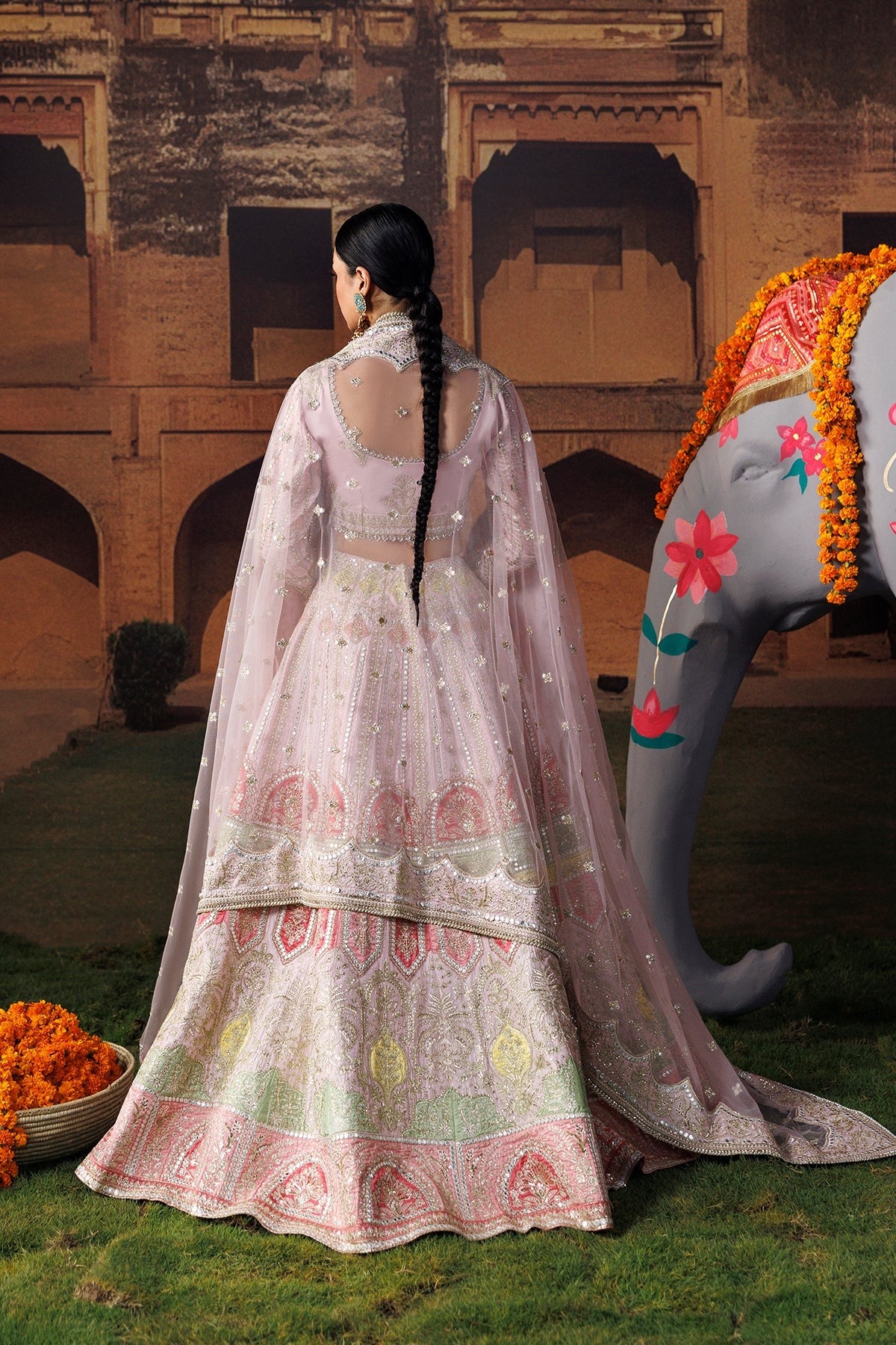 A woman wearing a light pink embroidered traditional Indian outfit stands with her back to the camera in front of a historic stone building, with a decorative elephant statue and orange flower garlands nearby.