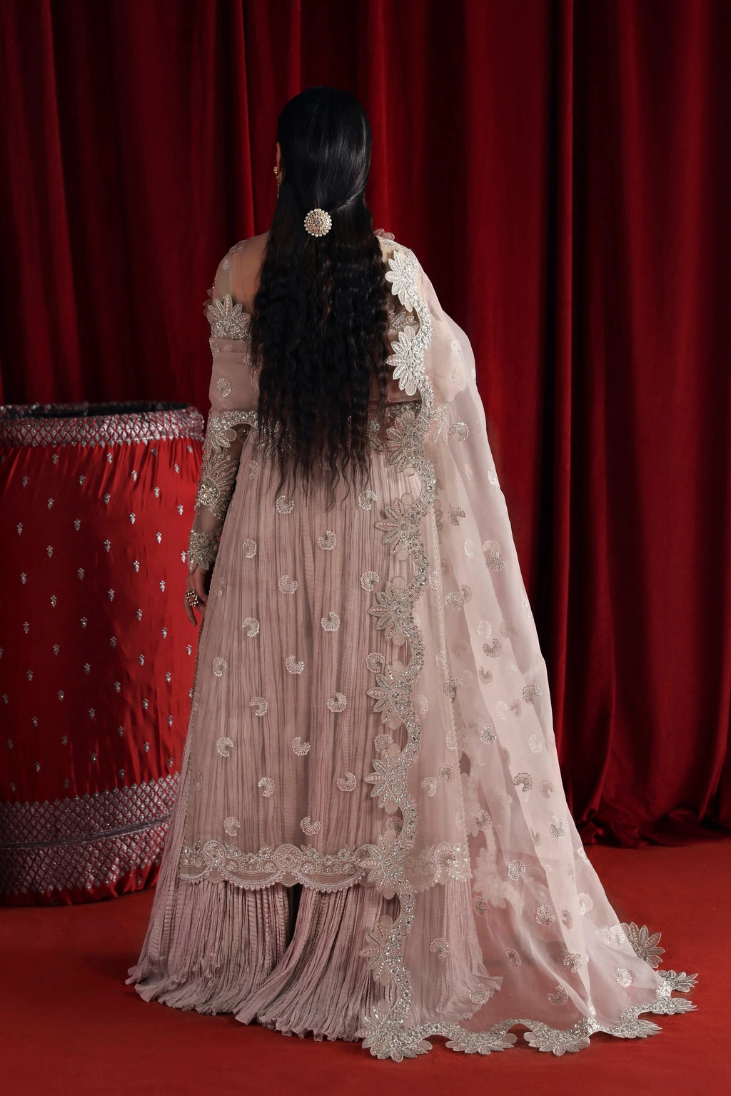 A woman viewed from behind wearing a light blush pink traditional gown with intricate silver embroidery and a matching sheer dupatta, set against a dark red curtain backdrop.