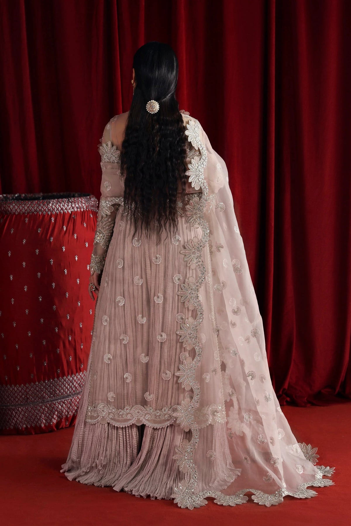 A woman viewed from behind wearing a light blush pink traditional gown with intricate silver embroidery and a matching sheer dupatta, set against a dark red curtain backdrop.
