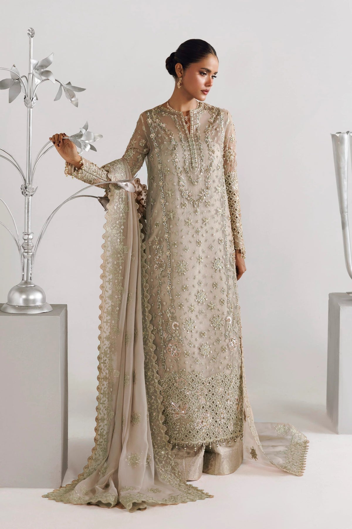 A woman models a traditional long beige embroidered kurta with matching dupatta and trousers in a studio setting with decorative silver props.