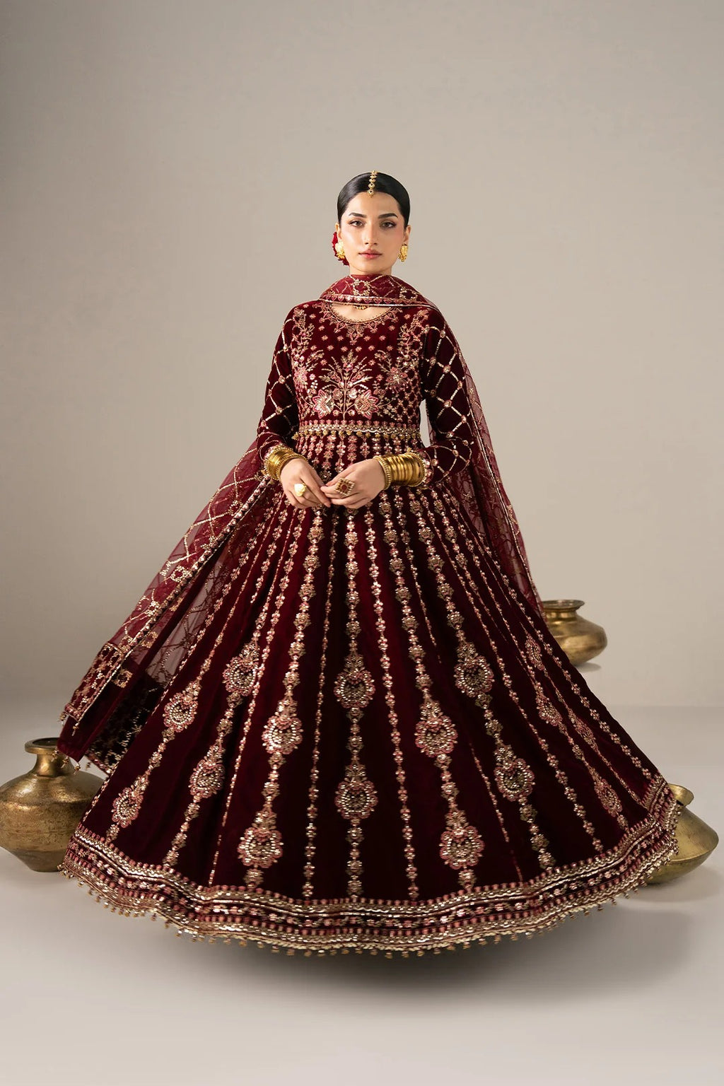 A woman modeling a traditional Indian bridal lehenga choli set in deep maroon velvet with intricate gold embroidery, a matching dupatta, and gold jewelry, posed against a neutral studio backdrop.