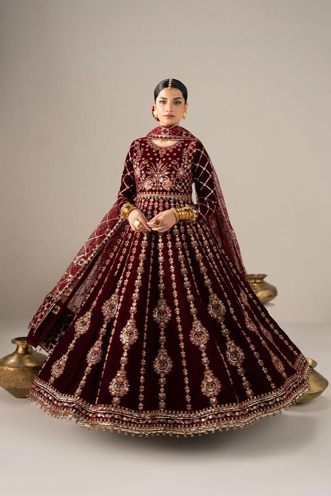 A woman modeling a traditional Indian bridal lehenga choli set in deep maroon velvet with intricate gold embroidery, a matching dupatta, and gold jewelry, posed against a neutral studio backdrop.