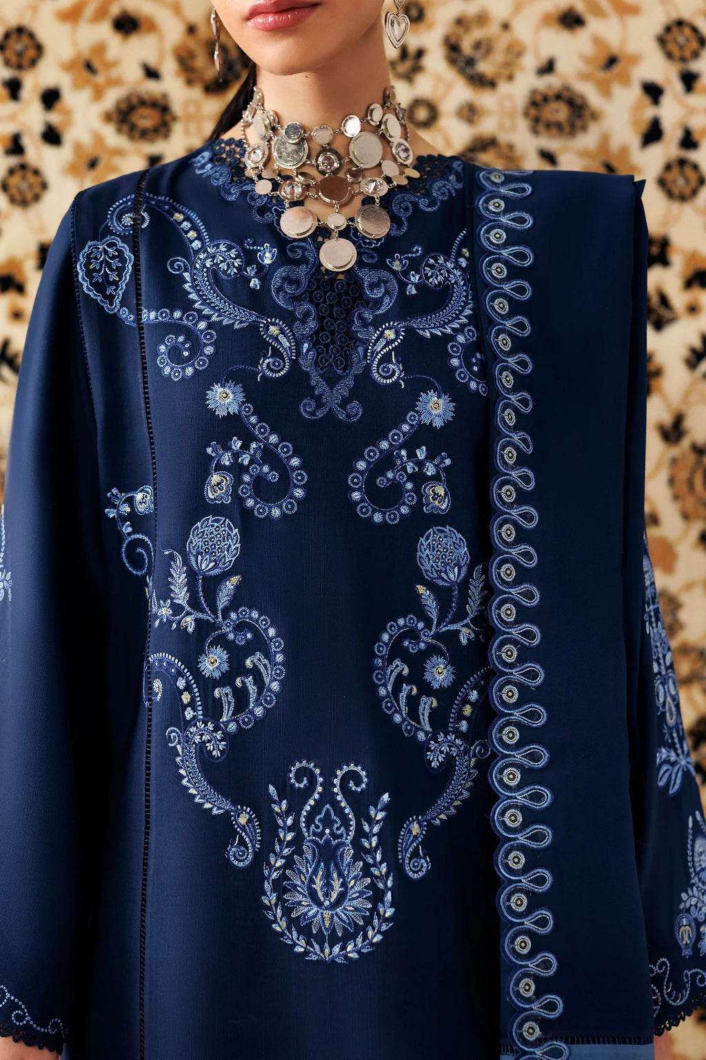 A close-up of a model wearing a navy blue embroidered tunic or kurta with intricate light blue and white floral patterns, paired with a chunky silver coin necklace, set against a patterned beige and black background.