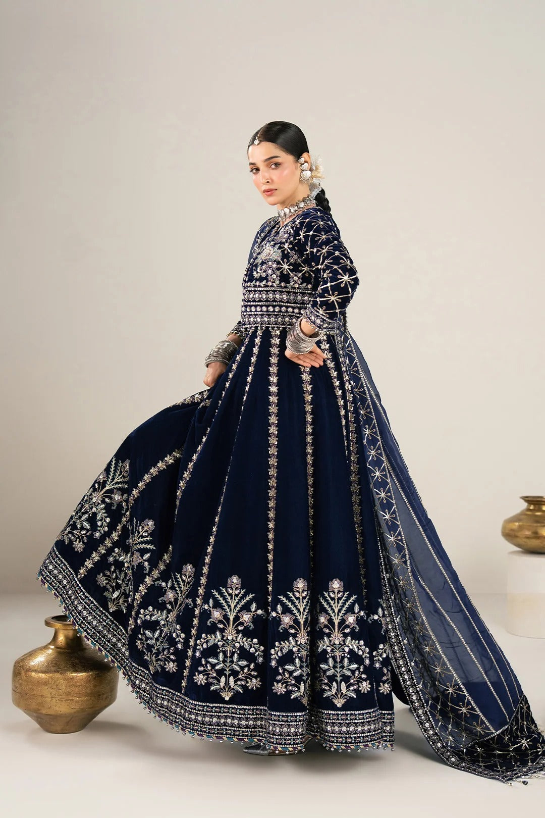 A woman models a navy blue traditional gown with intricate silver embroidery, featuring a high-necked bodice, full flared skirt, and matching dupatta, posed in a studio setting with decorative ceramic vases.