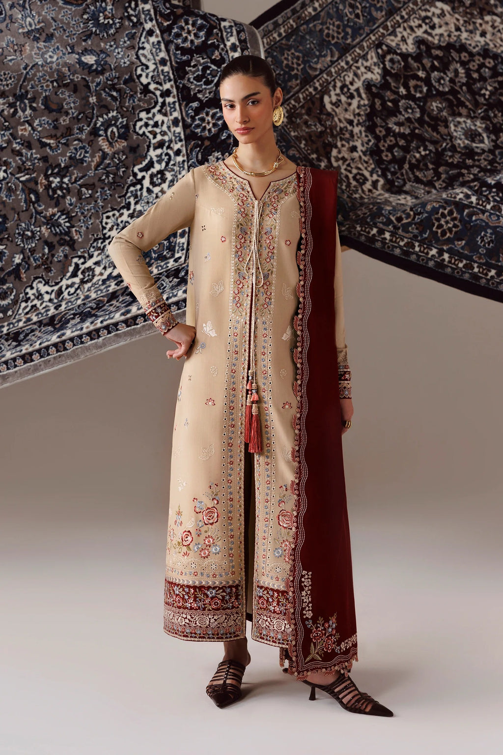 A woman models a traditional beige embroidered kurta set with a deep maroon dupatta, standing against a backdrop of patterned rugs.
