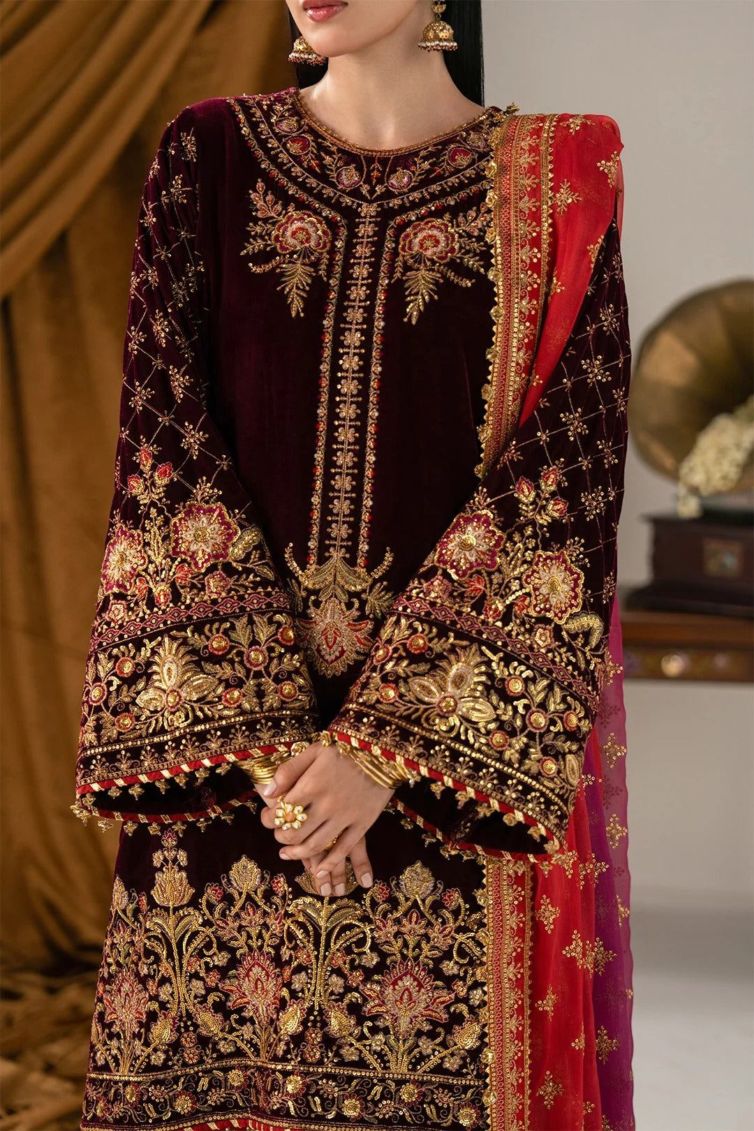 A close-up of a model wearing an ornate traditional South Asian outfit featuring a deep maroon velvet kurta heavily embroidered with gold and red floral patterns, paired with a matching red and gold dupatta.