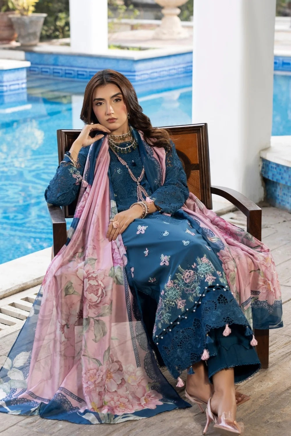 Woman in a blue embroidered outfit with a pink dupatta sitting by a pool.