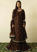 A woman modeling a dark brown traditional three-piece salwar kameez suit with white embroidered details, a matching dupatta, and a flared sharara skirt, set against a plain beige wall.