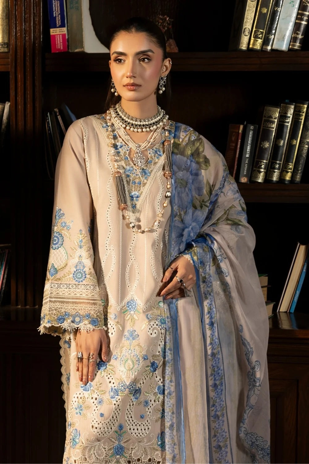 Woman in traditional embroidered outfit with jewelry in a room with bookshelves.
