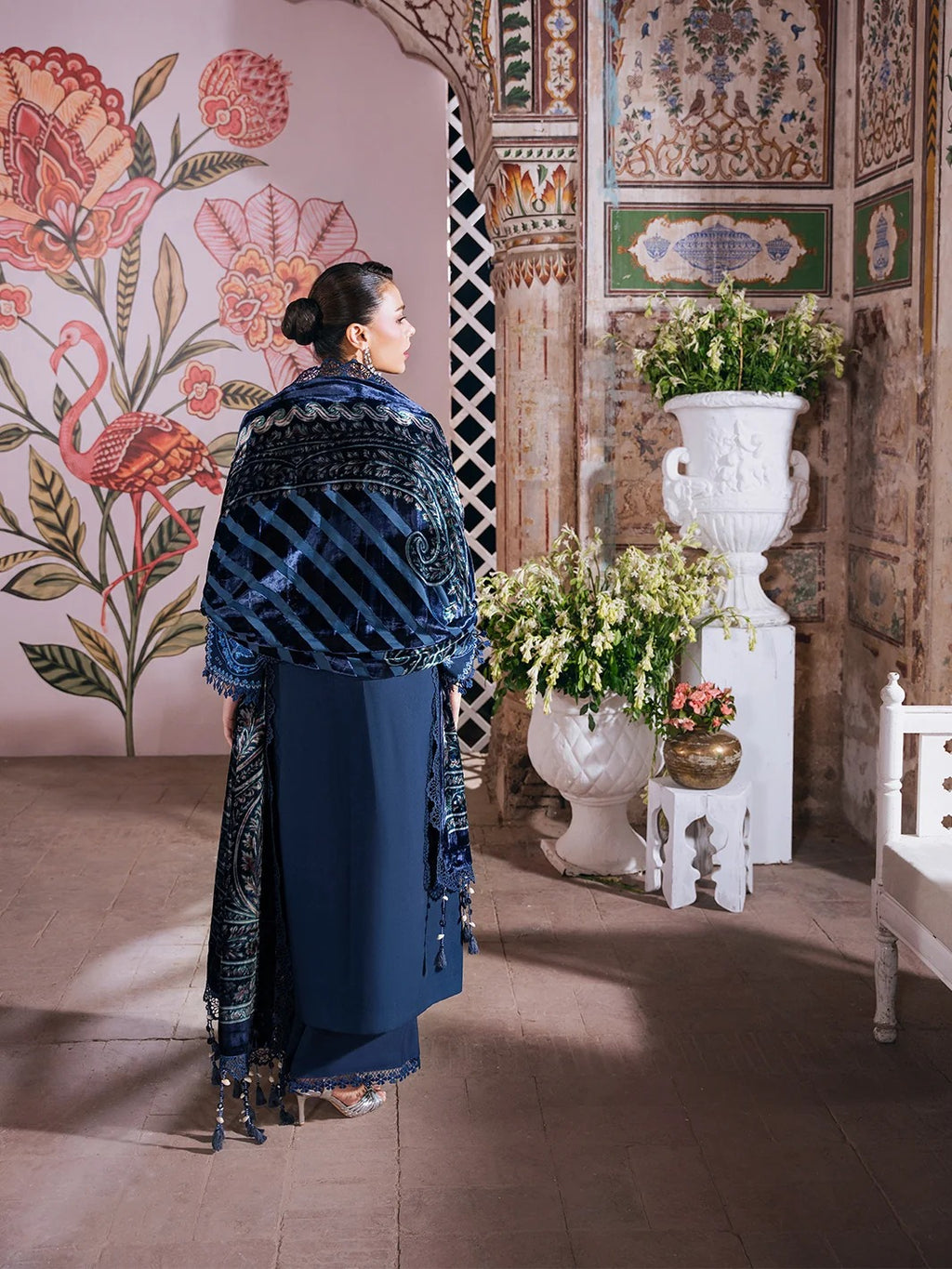 A woman wearing a dark blue traditional outfit with a patterned shawl stands in an ornate interior with floral wall murals, white planters, and warm stone flooring.