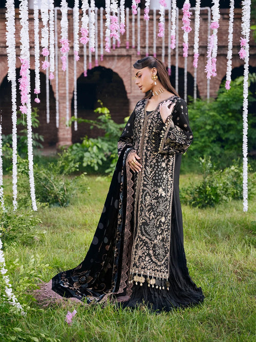 A woman stands outdoors in a grassy area wearing an ornate black traditional outfit with extensive gold embroidery and a matching sheer black dupatta. The background features hanging pink and white flower garlands and a brick archway.