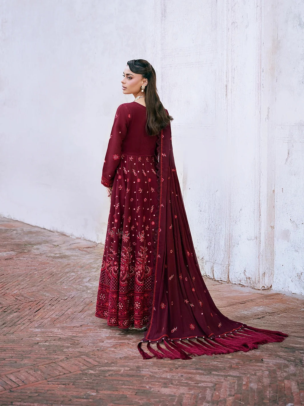 A woman stands in profile wearing a long maroon traditional outfit with a matching sheer dupatta and train, set against a white wall and brick floor.