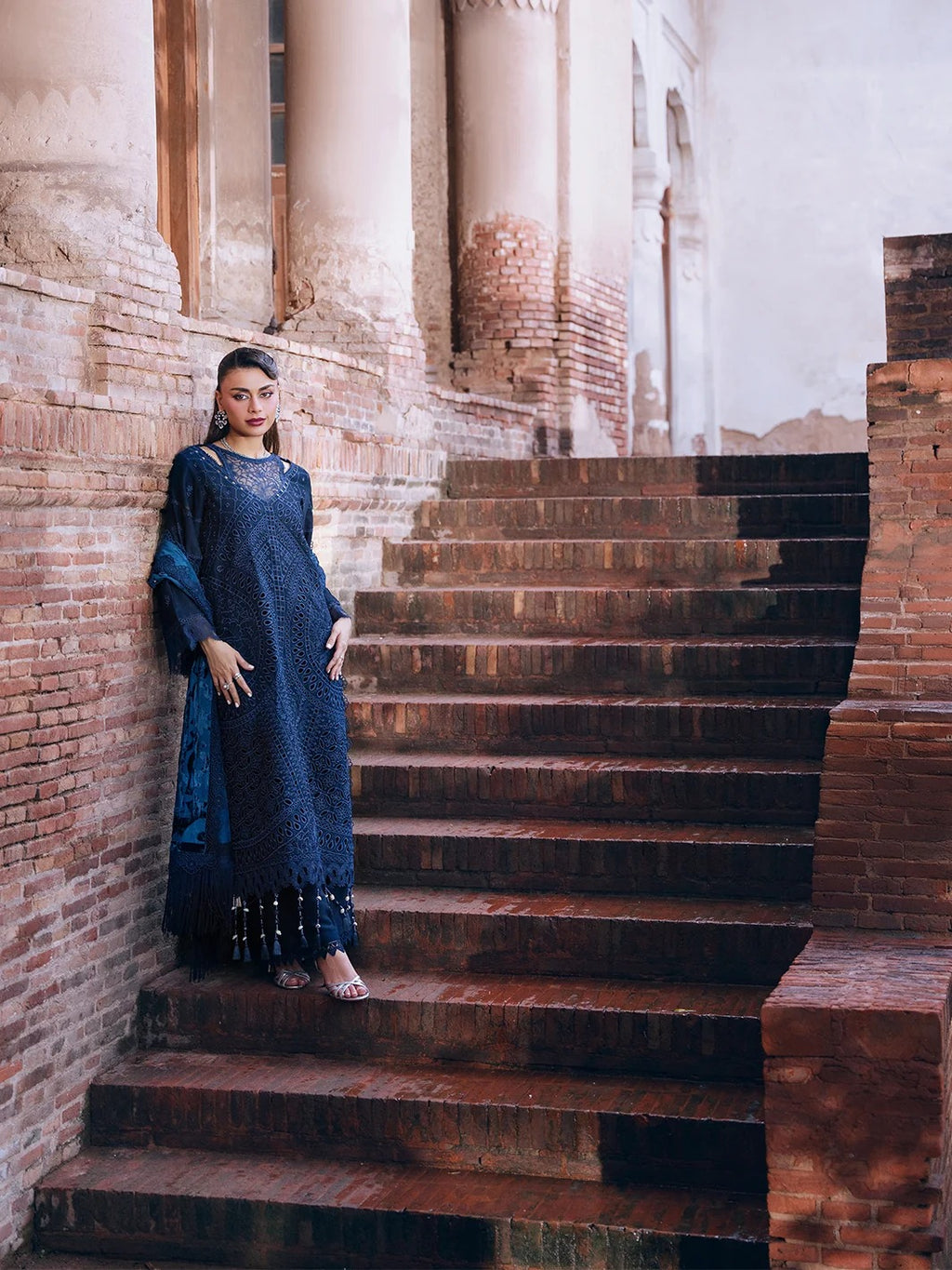 A woman wearing a dark navy blue embroidered kurta and matching dupatta stands on stone stairs in front of a brick and plaster building with columns.