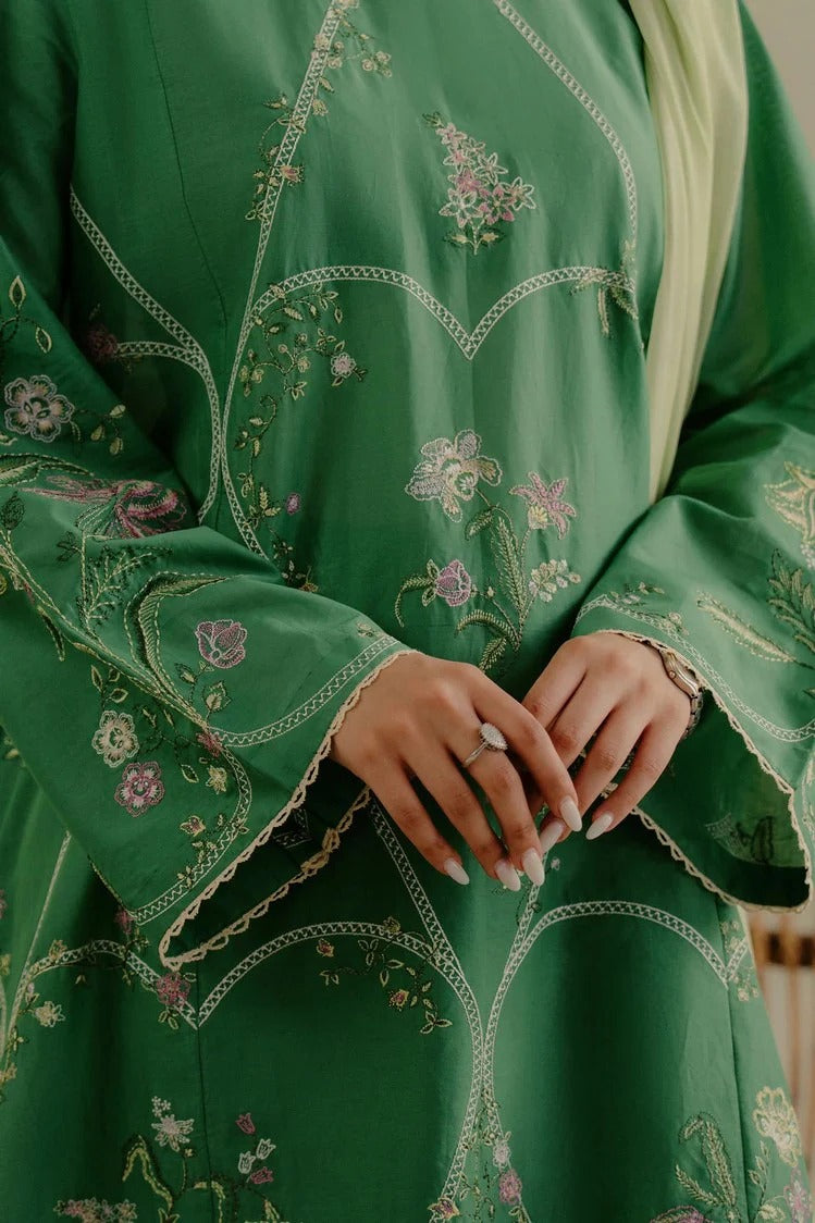 A close-up of a person wearing a green embroidered traditional garment with floral detailing and lace-trimmed cuffs, with hands clasped in front.