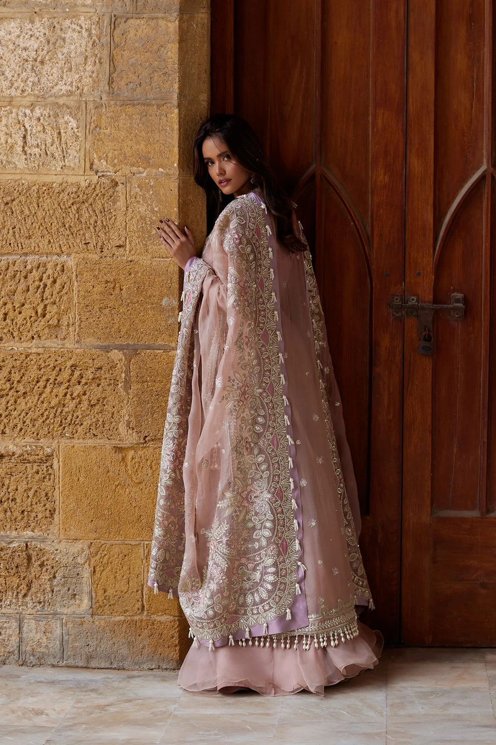 A woman wearing a dusty pink traditional outfit with intricate gold embroidery stands in profile between a textured stone wall and a dark wooden door.