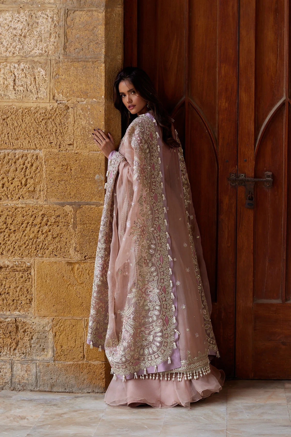 A woman wearing a dusty pink traditional outfit with intricate gold embroidery stands in profile between a textured stone wall and a dark wooden door.