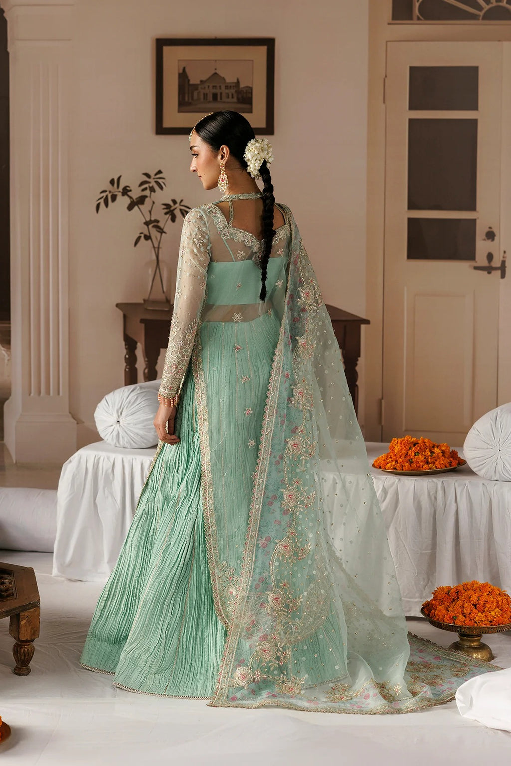 A woman wearing a mint green traditional Indian lehenga choli set with a matching sheer embroidered dupatta, standing in a decorated indoor room.
